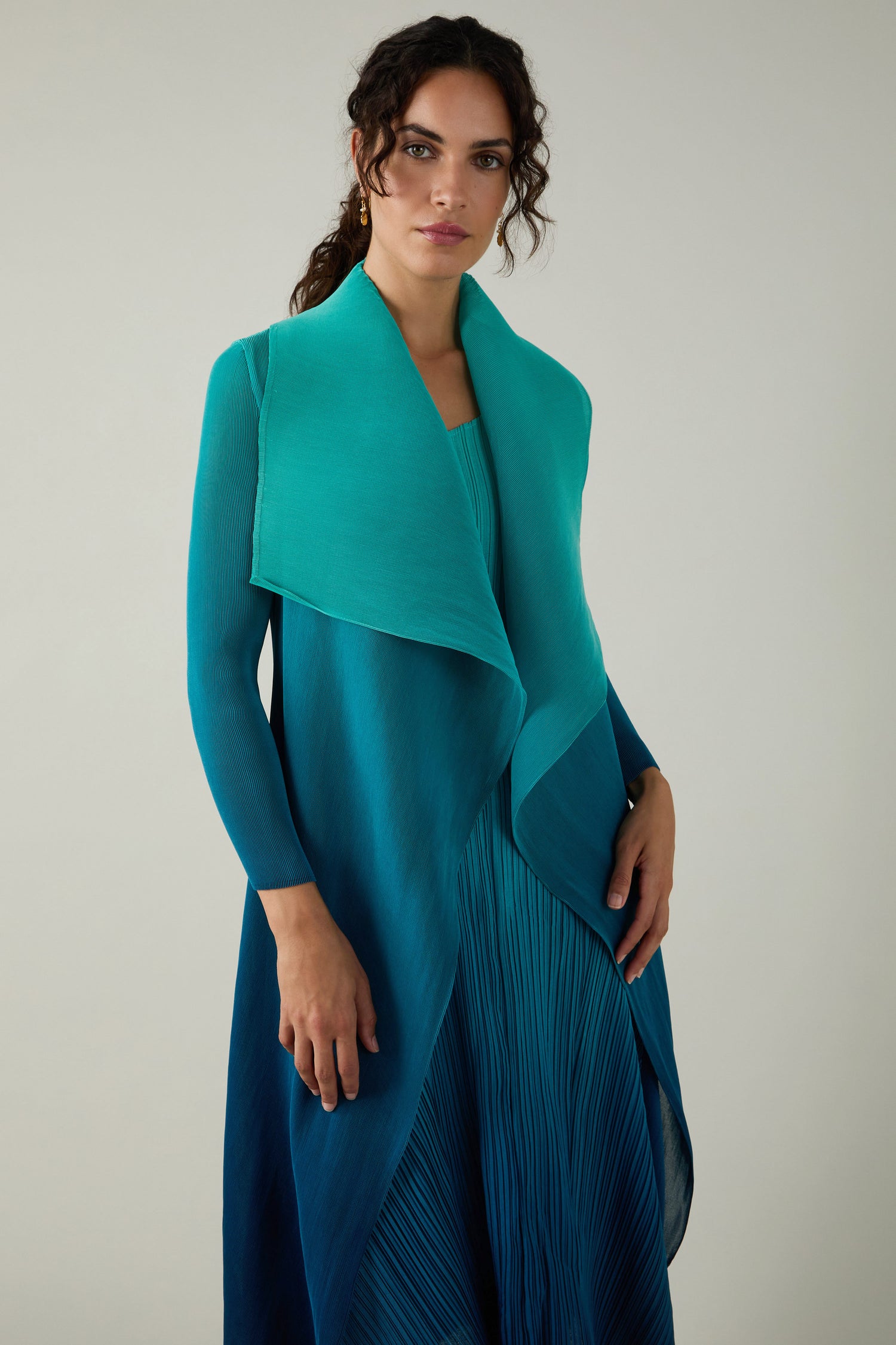 Woman standing and facing forward, wearing an Alquema Pleated Jacket: a finely pleated, gradient blue-to-teal jacket with a draped, asymmetrical collar inspired by Japanese shibori techniques.
