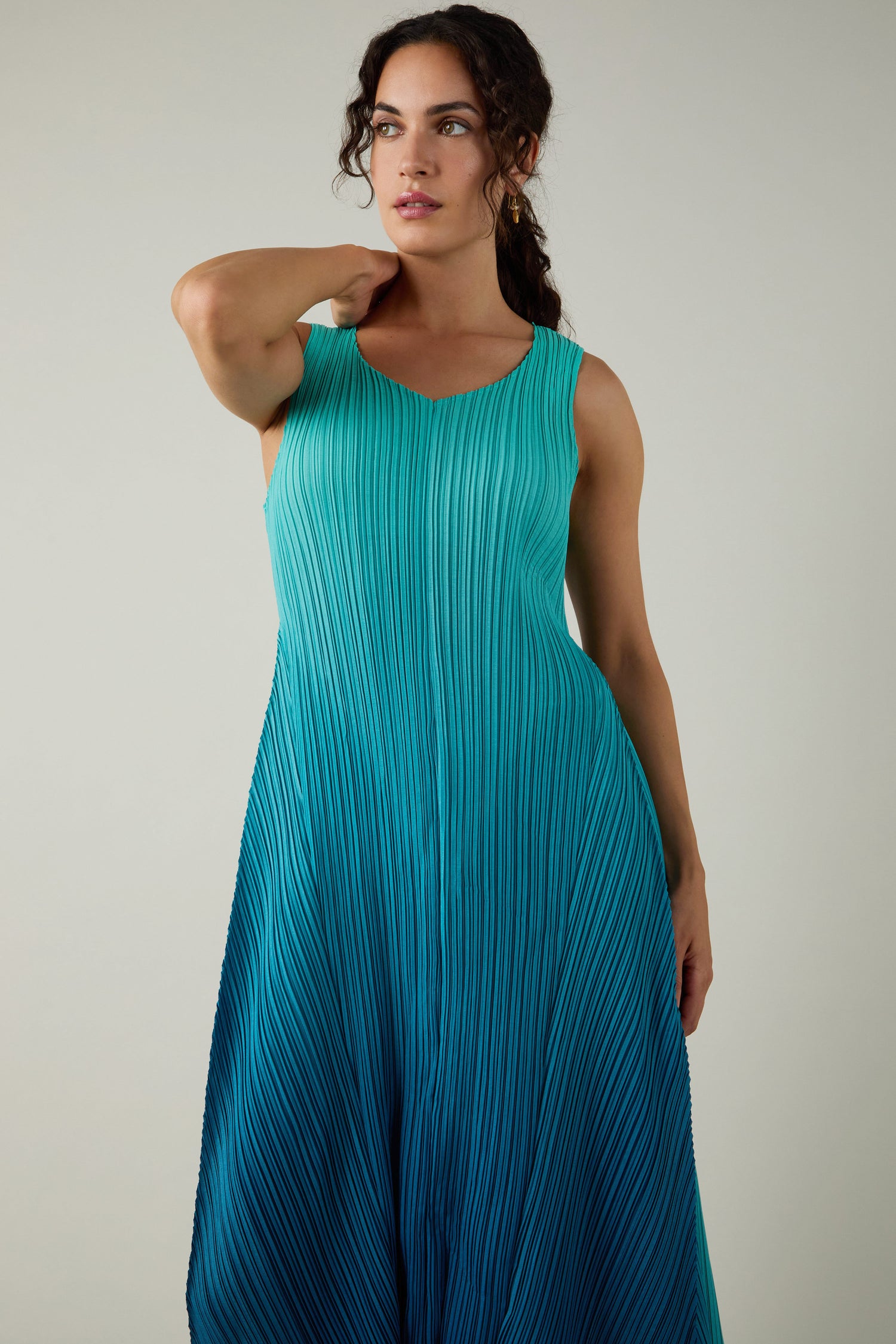 A woman stands against a plain light background, wearing the Pleated Dress in teal and blue with shibori pleating, touching her neck with one hand.