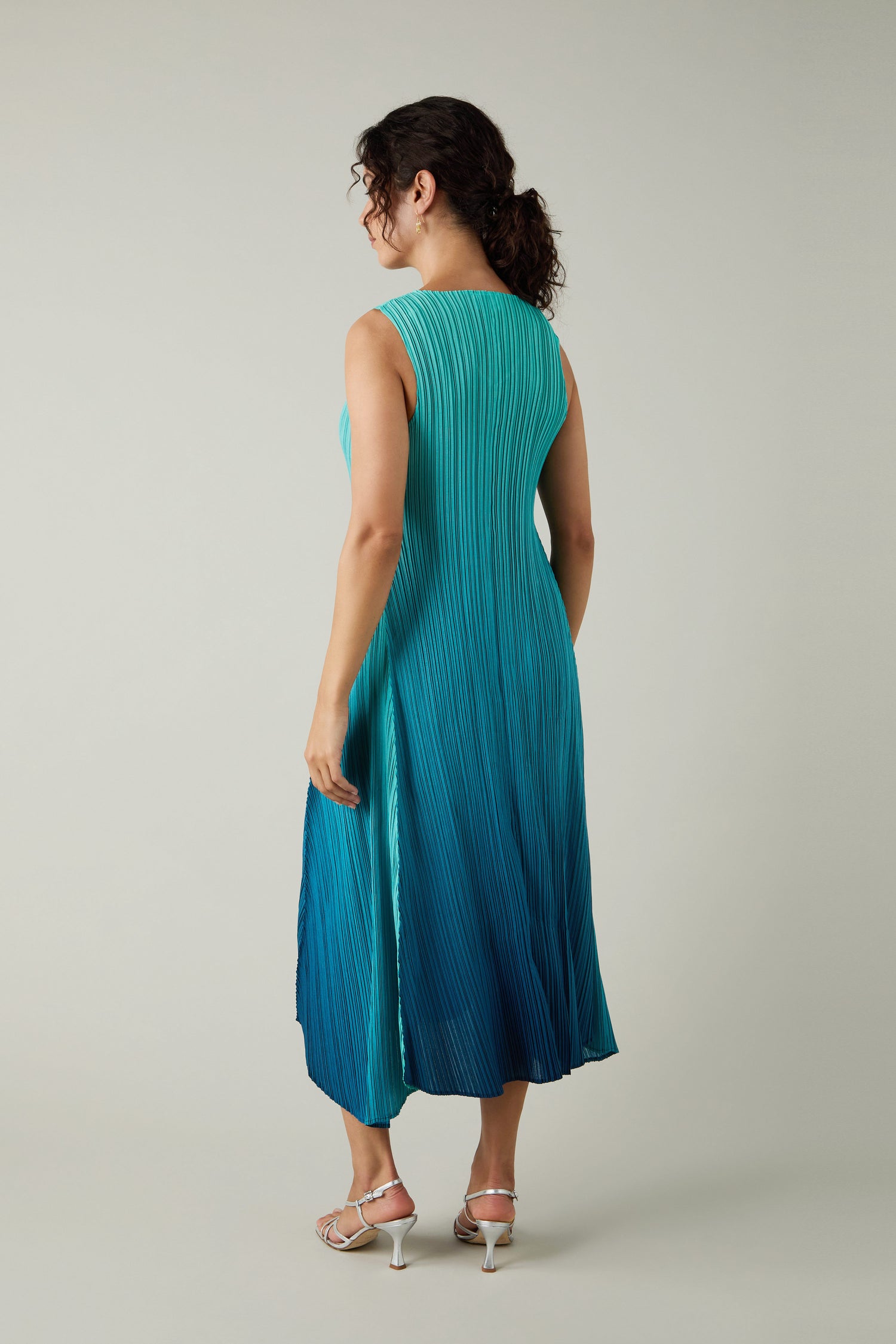 A woman faces away wearing a Pleated Dress, featuring shibori pleating in a sleeveless teal-to-blue gradient, paired with white heeled sandals against a plain light background.