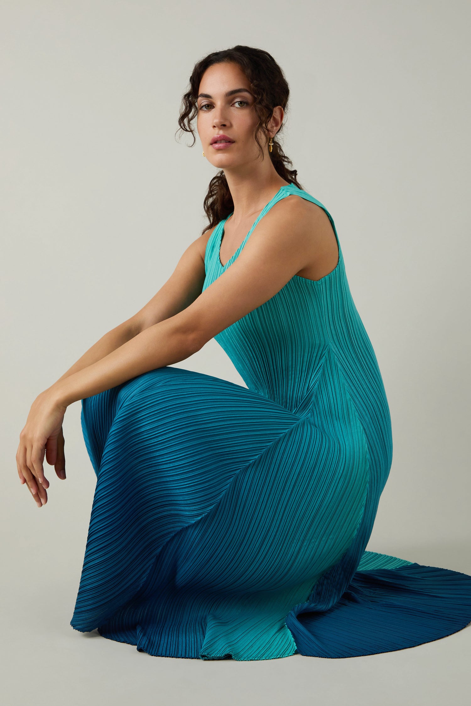A woman with dark hair wears a Pleated Dress, showcasing teal and blue shibori pleating, as she sits on the floor against a plain light background.