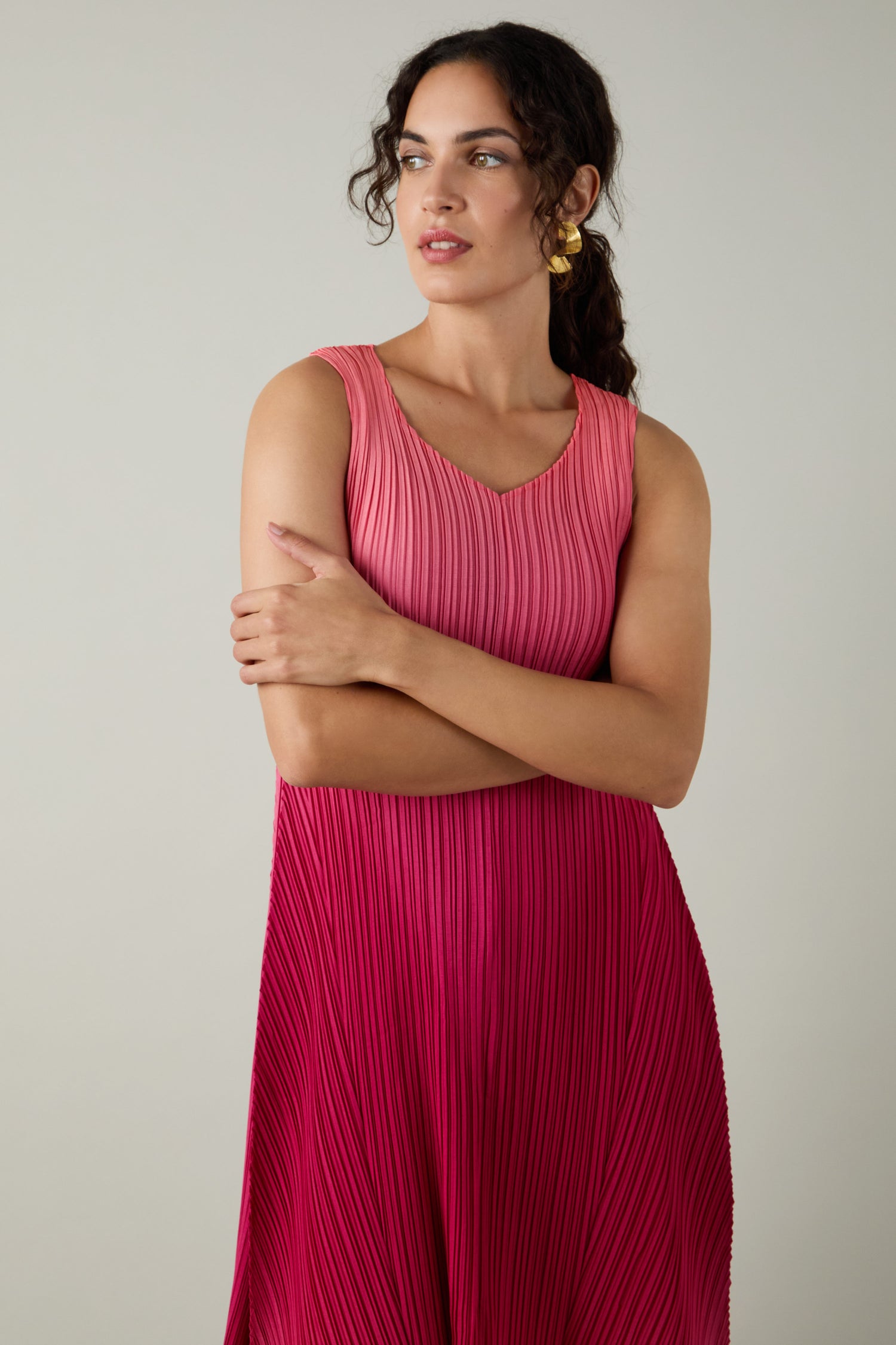 A woman with wavy dark hair and gold hoop earrings wears a Pleated Dress featuring shibori pleating in a sleeveless pink-to-fuchsia ombre design, standing with arms crossed against a plain background.
