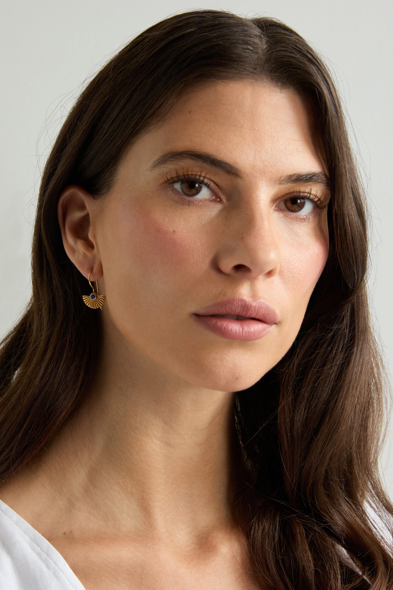 A woman with long brown hair, wearing a white top and the Gold Vermeil Lapis Fan Earrings, gazes slightly to the side against a plain background.