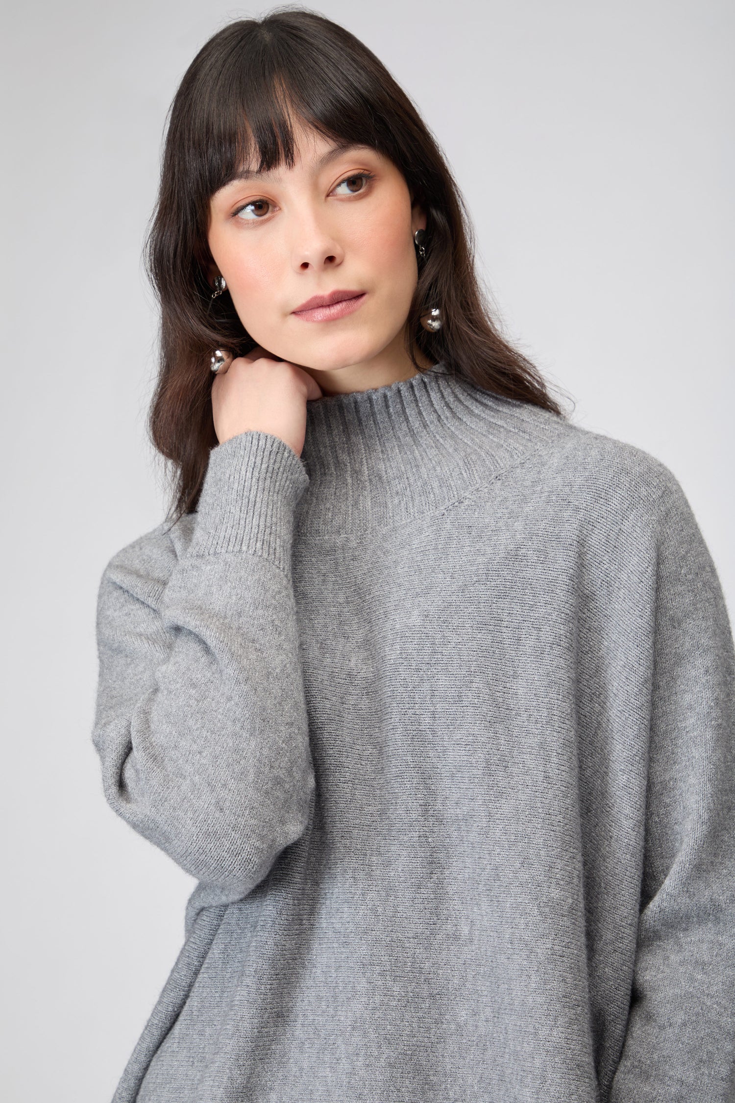 A woman with long brown hair wears the Boxy High Neck Knit in gray, looking slightly to the side with a neutral expression.