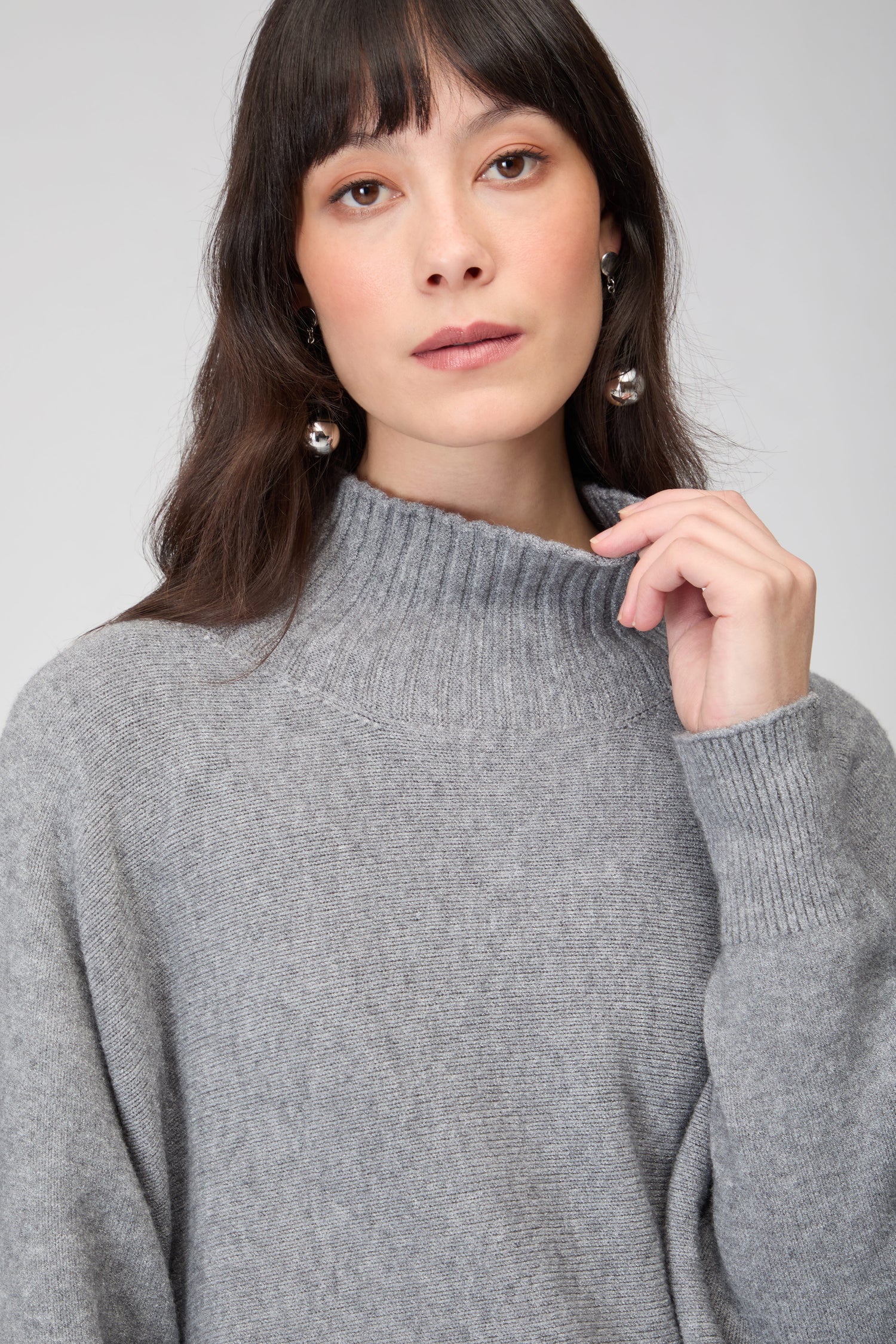 A woman with long brown hair wears the Boxy High Neck Knit in grey and silver earrings, posing against a plain background.