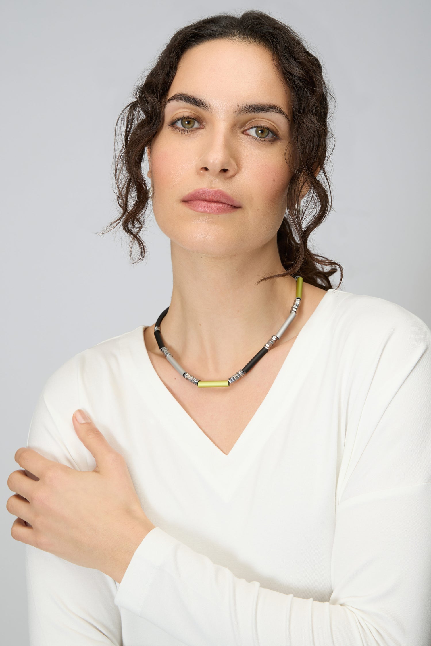 Woman with wavy brown hair wearing a white V-neck top and the Atomic Silver Aluminium Hematite Necklace, looking at the camera with a neutral expression.