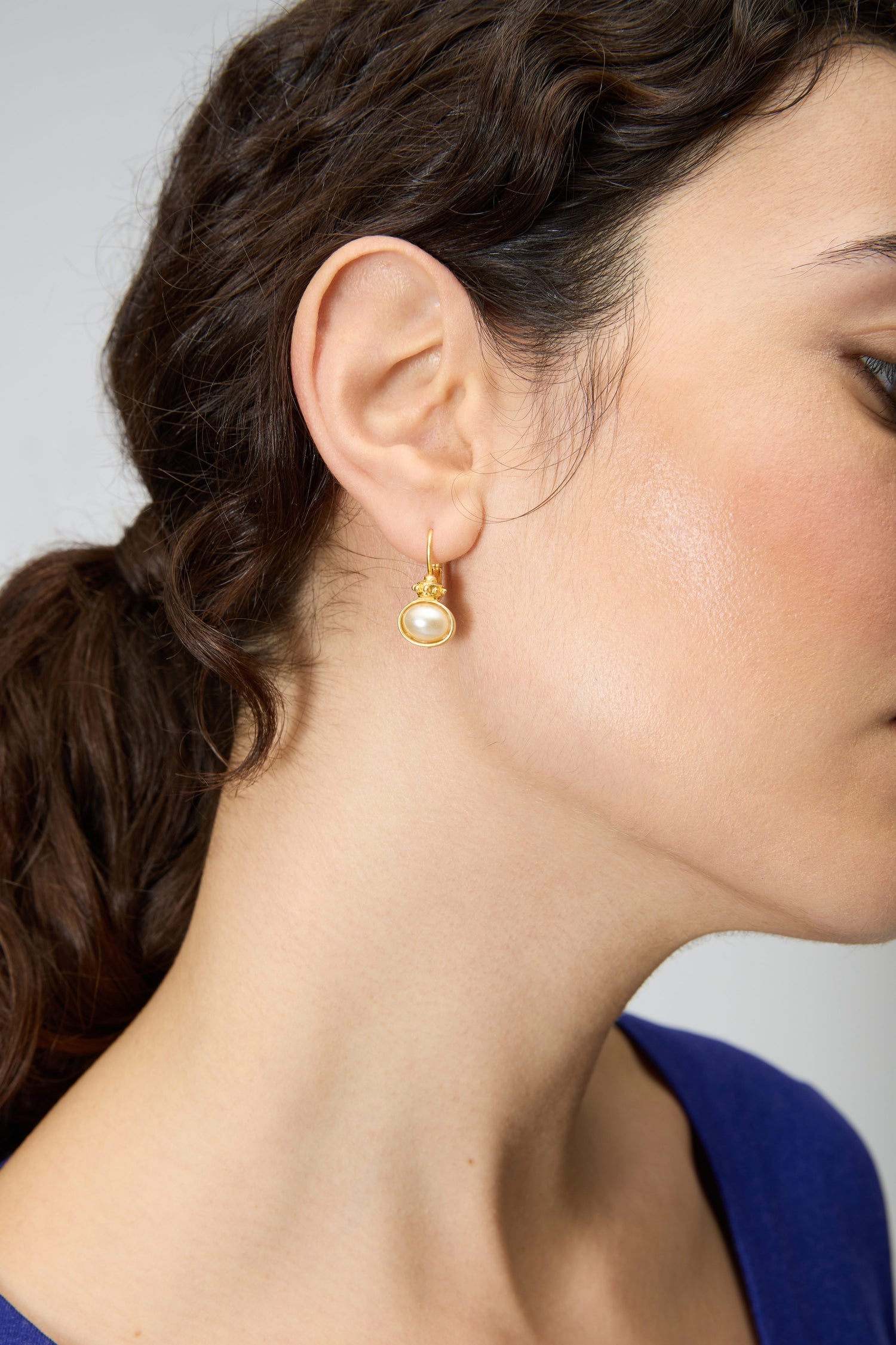 Close-up of a woman's ear wearing Gold Plated Pearl Drop Earrings; her dark hair is tied back, and she is in a blue top.