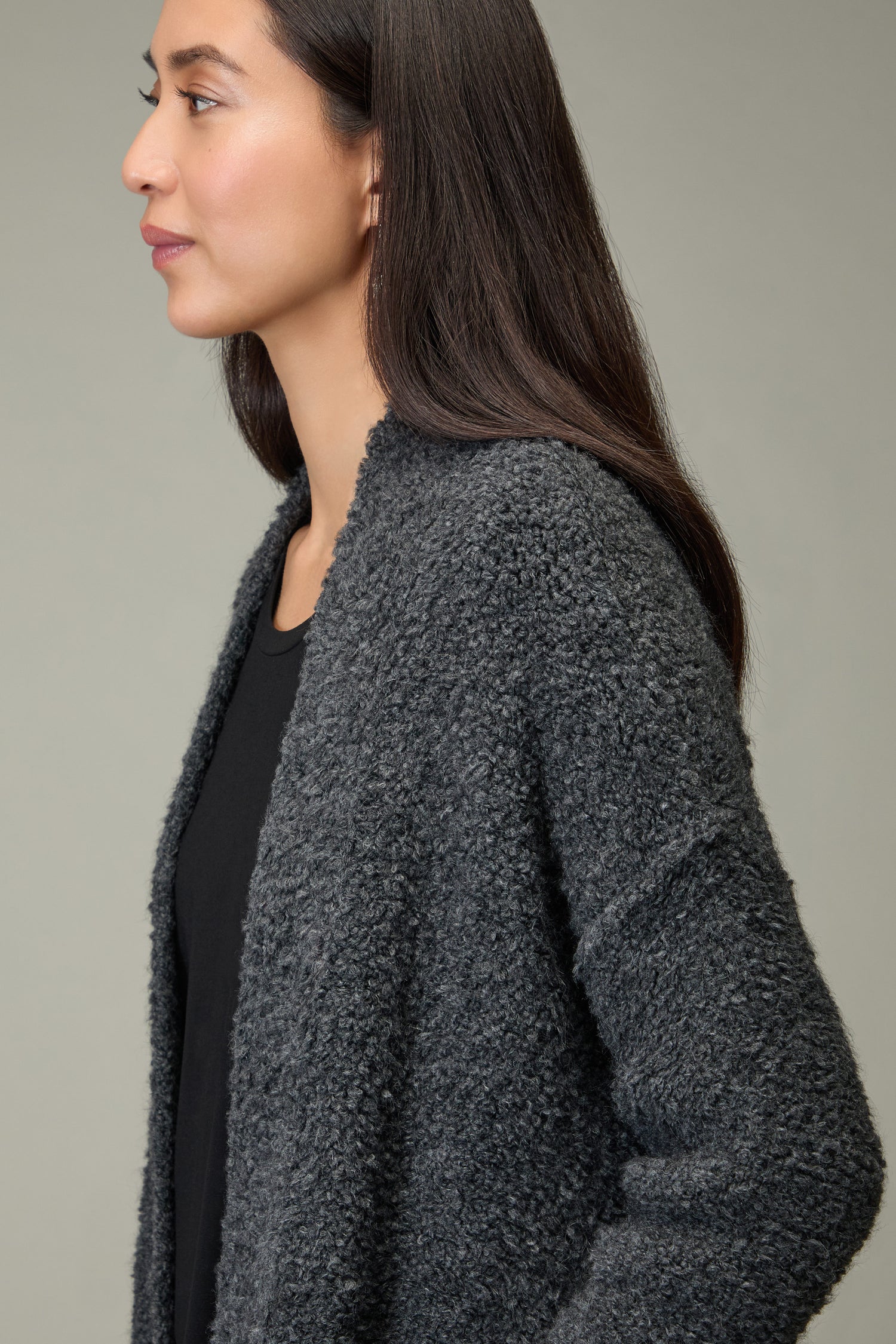 Woman with long dark hair wears the Charcoal Boucle Knit Long Cardi and a black top, facing left against a plain light gray background—an ideal choice to elevate your autumn wardrobe.