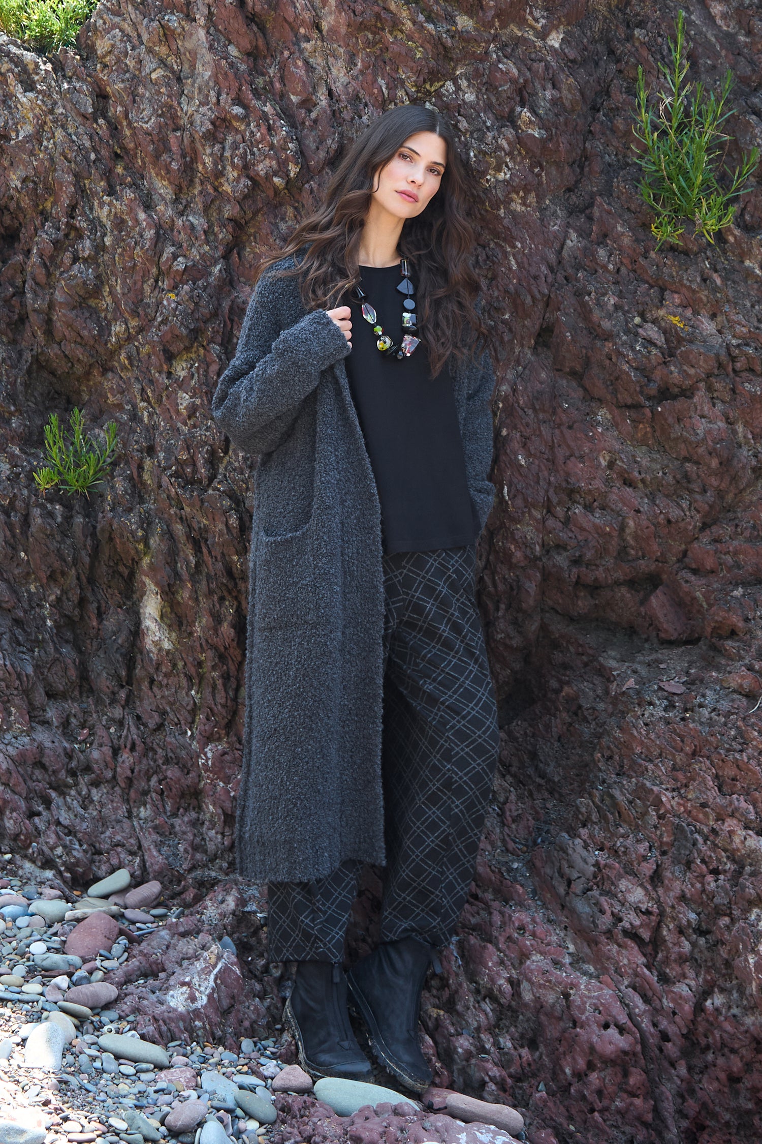 A woman stands against a rocky wall, wearing the Charcoal Boucle Knit Long Cardi (GOK5065-BOK), black patterned pants, black boots, and a colorful beaded necklace.