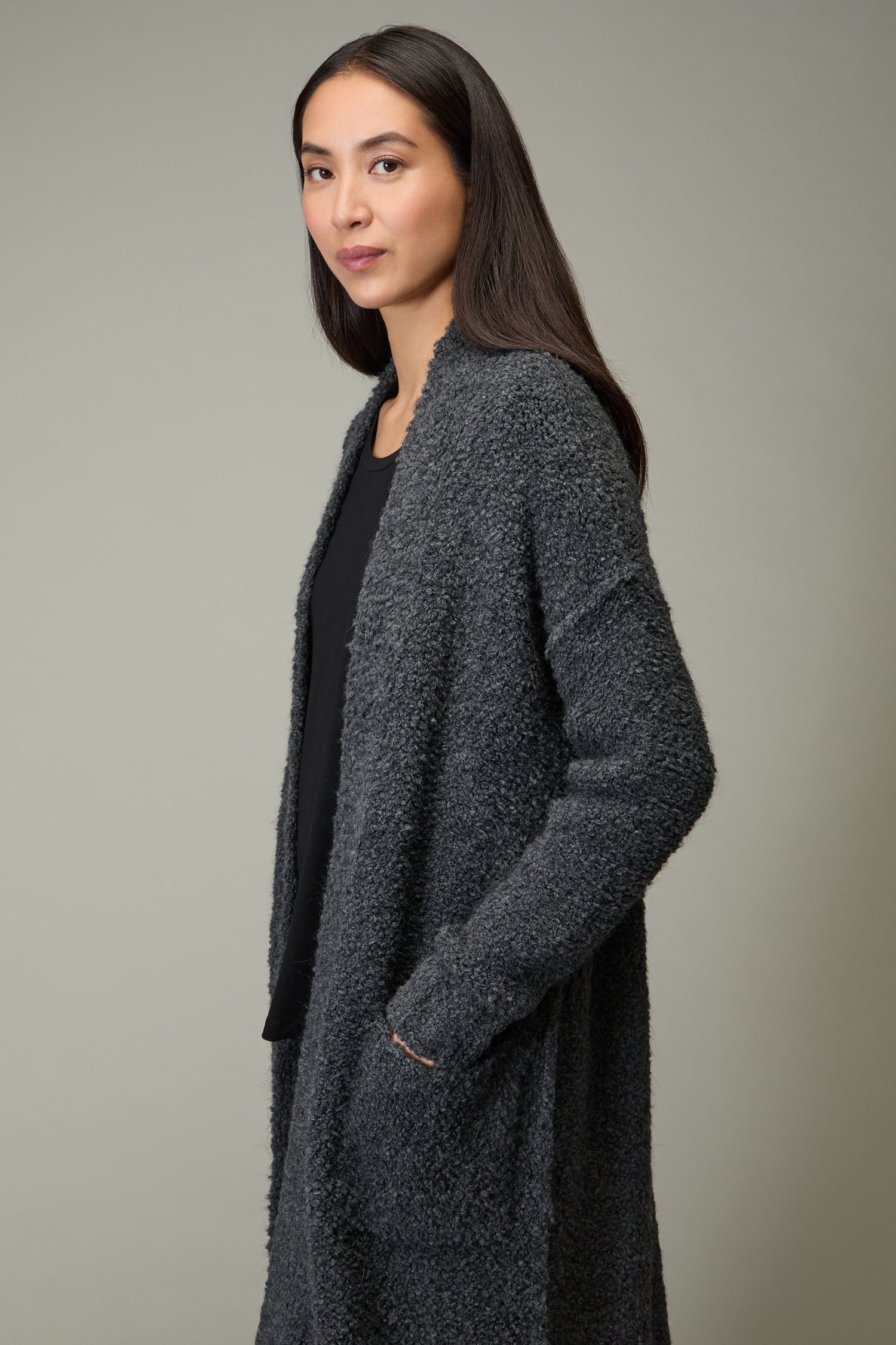 A woman with long dark hair stands slightly to the side, hand in pocket, wearing the Charcoal Boucle Knit Long Cardi over a black top—an effortless staple for any autumn wardrobe.