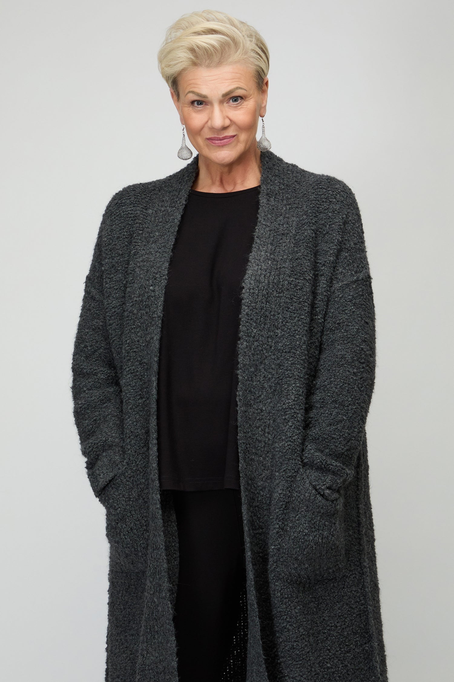 Older woman with short gray hair wears the Charcoal Boucle Knit Long Cardi over a black shirt, standing against a plain light background with hands in pockets—ideal for autumn layering.