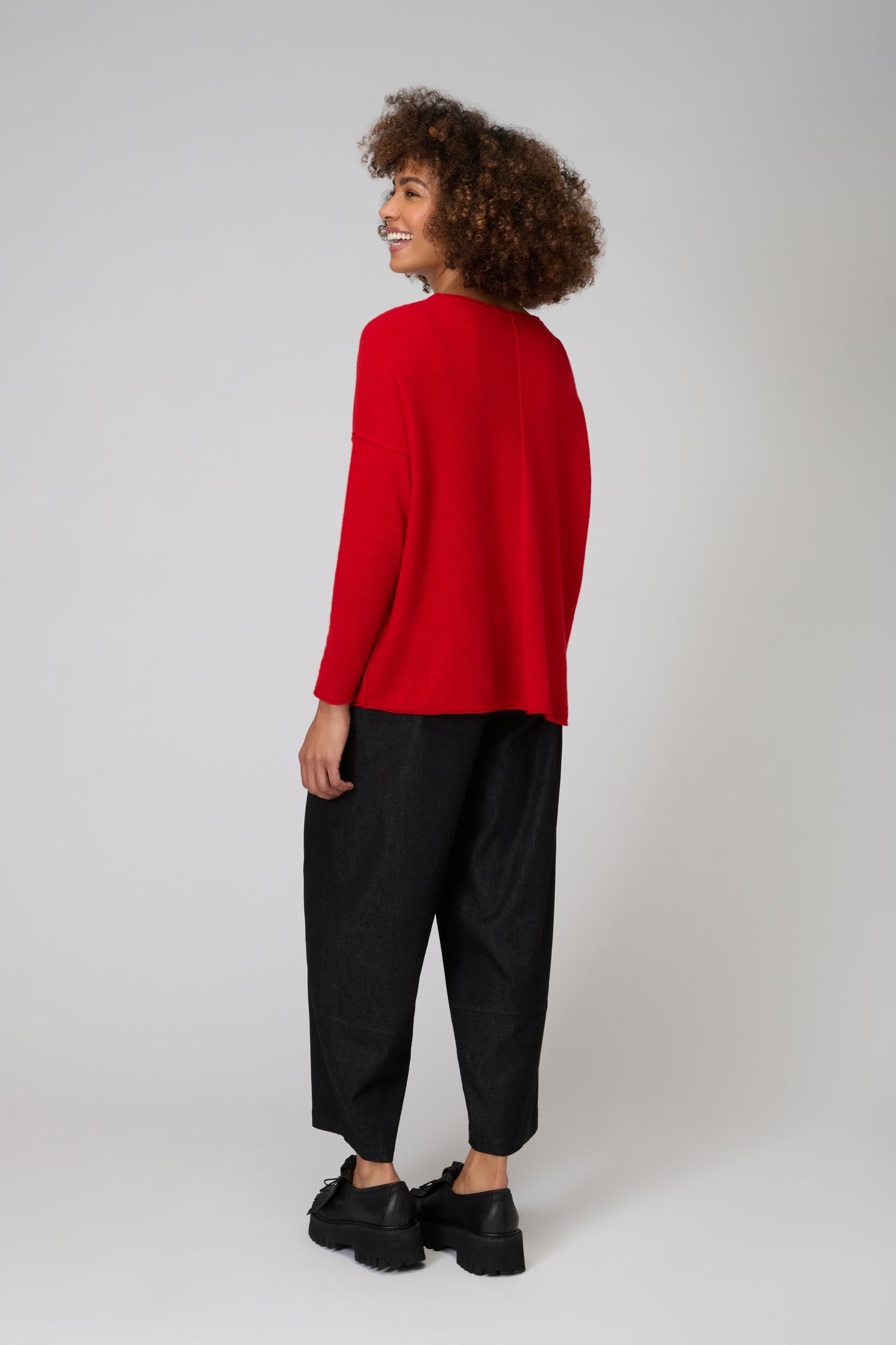 A person faces away, wearing a loose Merino Pocket Jumper in red, black wide-leg pants, and black shoes against a plain light background.
