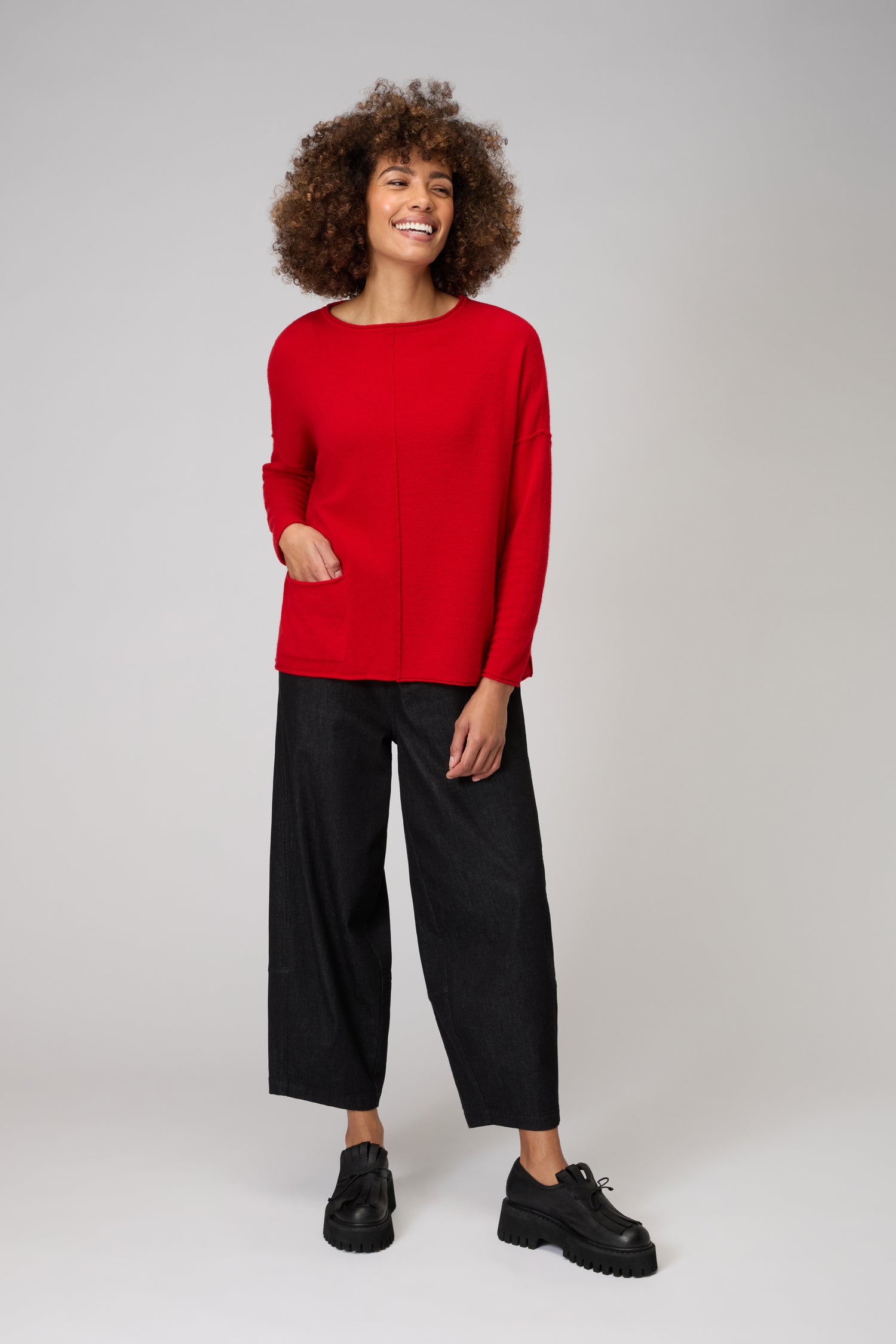 A person stands against a light gray background wearing the red Merino Pocket Jumper, black wide-leg pants, and black platform shoes, smiling with one hand in their pocket.