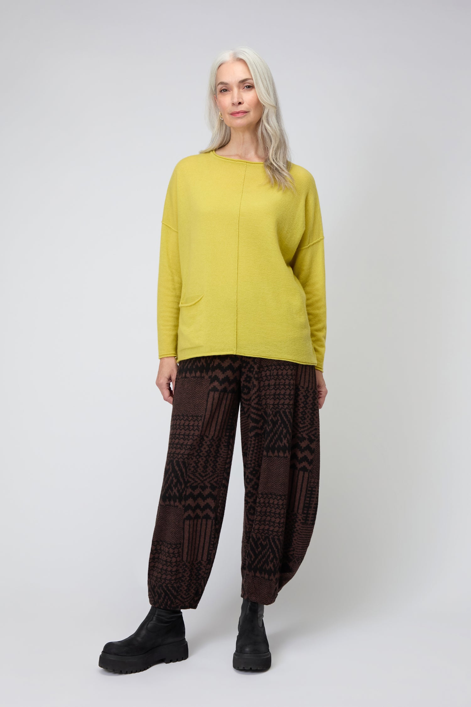 A woman with long gray hair wears the yellow Merino Pocket Jumper, patterned brown wide-leg pants, and black boots while standing against a plain light background.