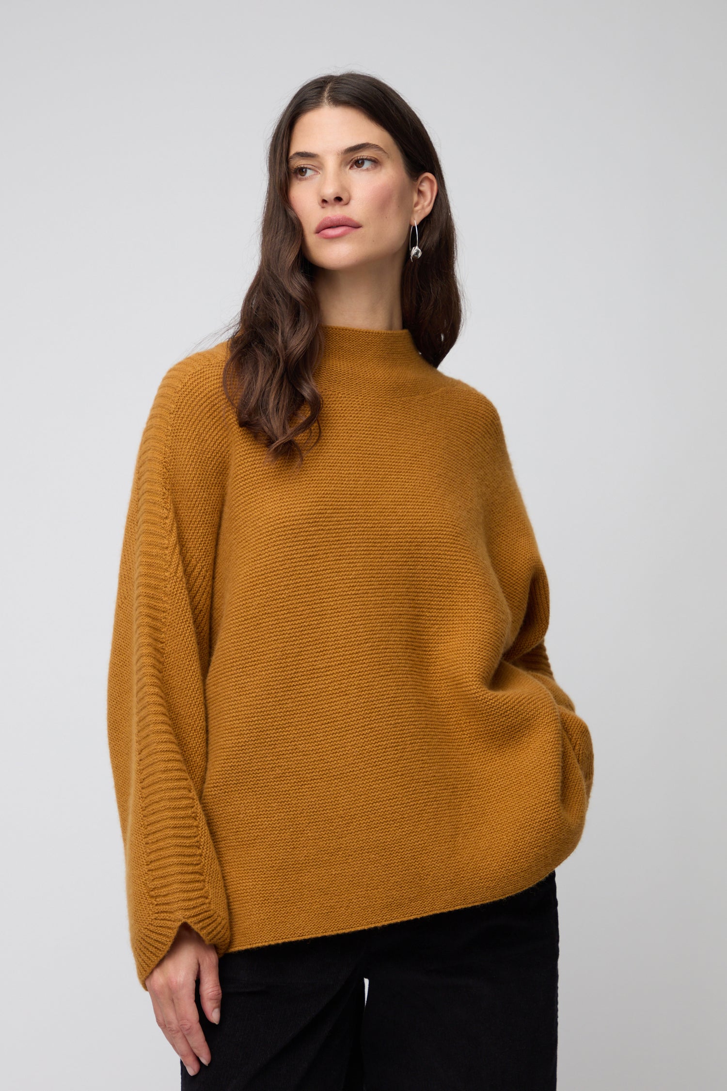 A woman faces forward against a plain light gray background, wearing the Merino Wool Stitch Detail Jumper in a relaxed boxy silhouette, paired with black pants and black shoes with white spots.