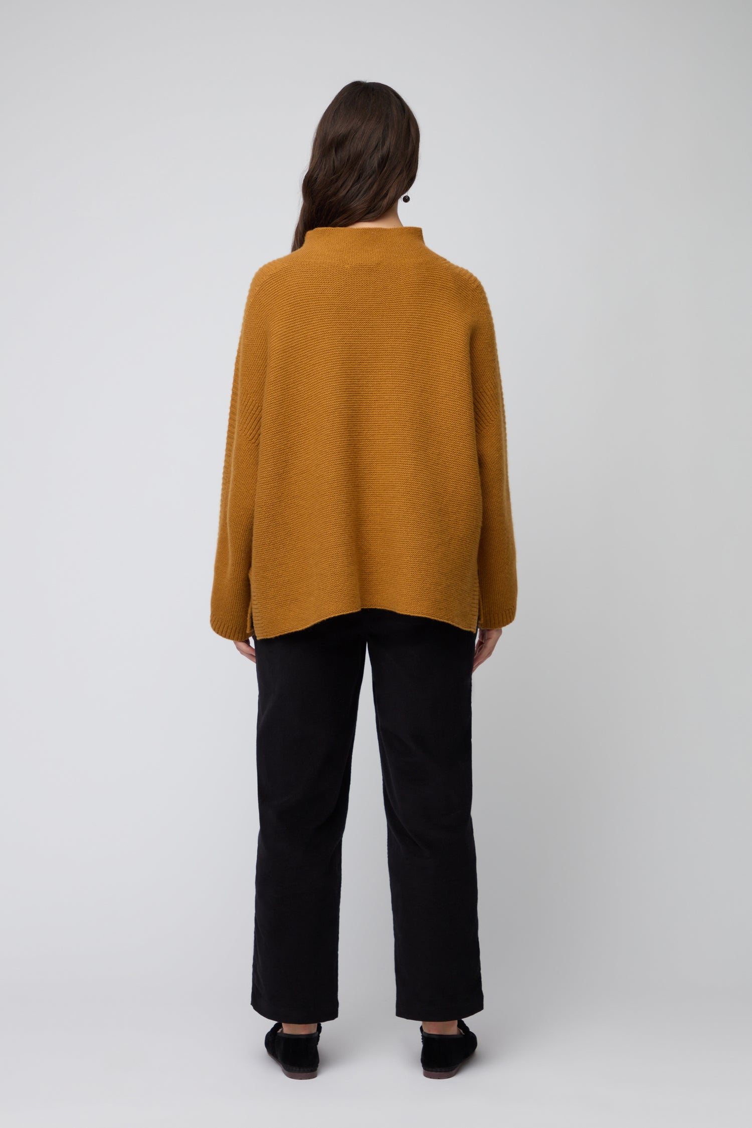 A person with long brown hair, wearing the Merino Wool Stitch Detail Jumper in mustard yellow and black pants, stands facing away from the camera against a plain light background.
