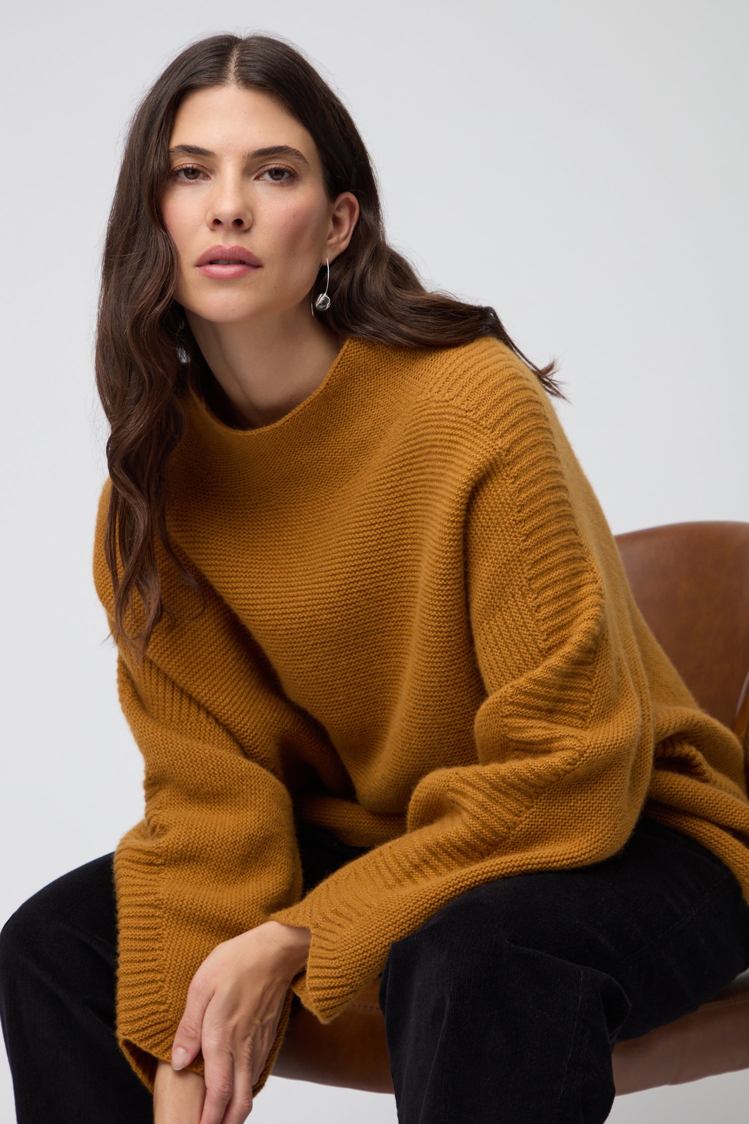 A woman with long dark hair wears the Merino Wool Stitch Detail Jumper in mustard, featuring a relaxed boxy silhouette. She stands against a plain light gray background, facing the camera.