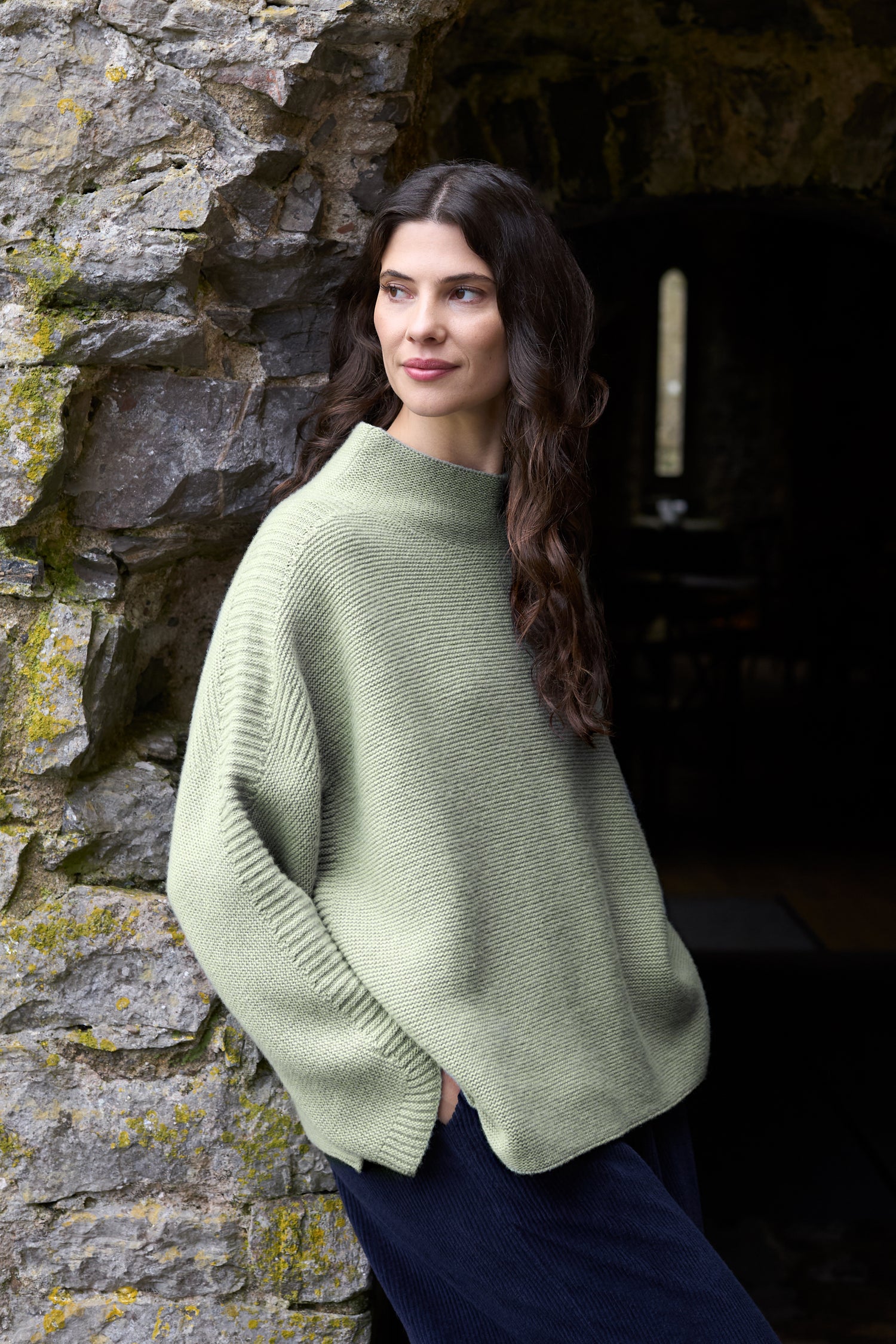 A woman with long dark hair wears the Merino Wool Stitch Detail Jumper in a relaxed, boxy fit and green color, standing against a stone wall, gazing to the side with her hands in her pockets.