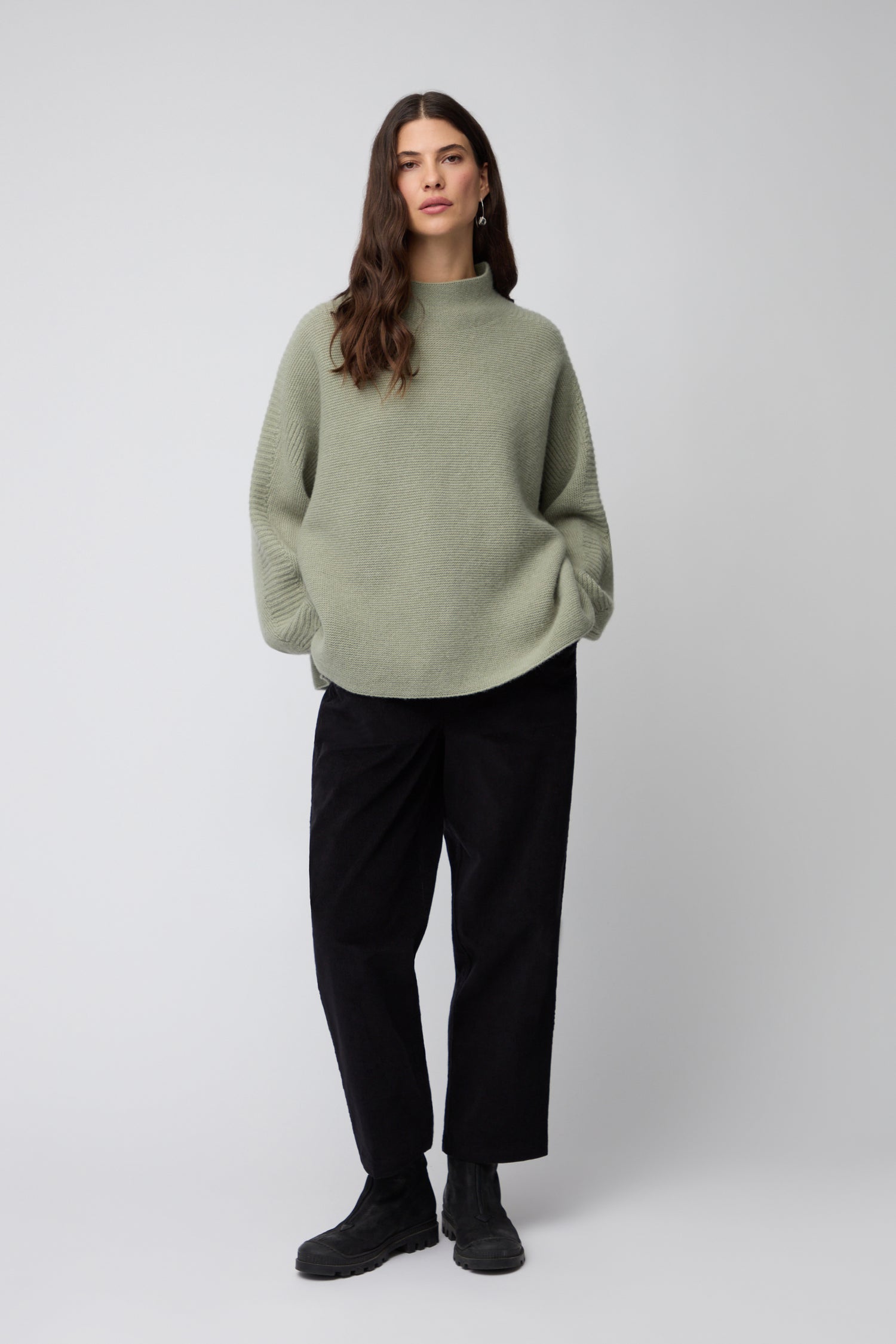 A person stands against a plain background wearing the Merino Wool Stitch Detail Jumper in sage green, paired with black pants and black shoes, hands in their pockets.