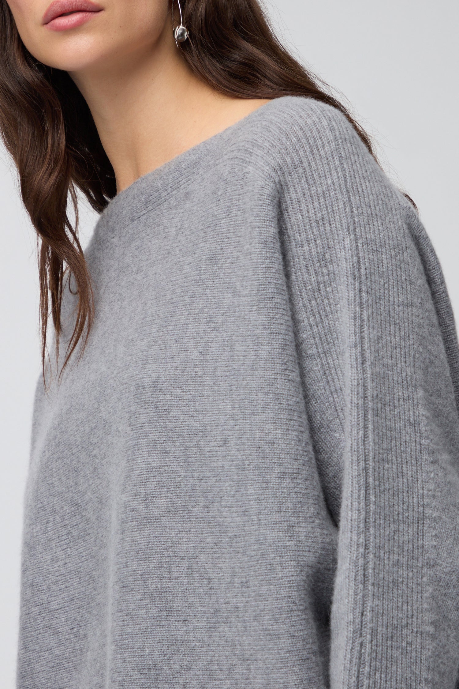 A woman wearing the Merino Rib Detail Jumper—a lightweight knit in light gray with an asymmetrical neckline. Only the lower half of her face and upper body are visible.