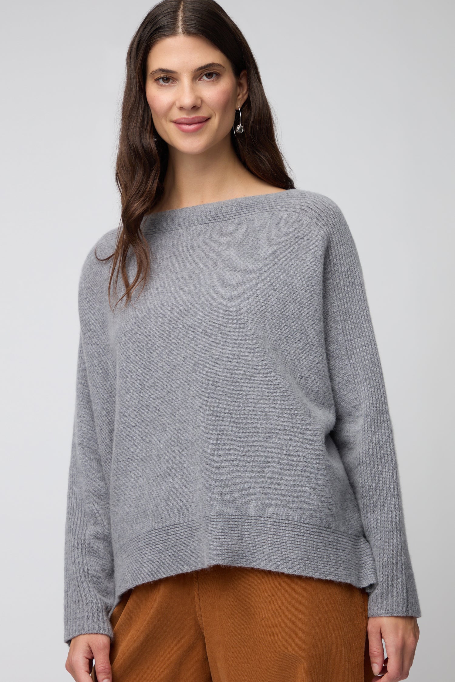 A woman with long dark hair wears the Merino Rib Detail Jumper, a loose-fitting gray knitwear piece, paired with brown pants, standing against a plain light gray background.