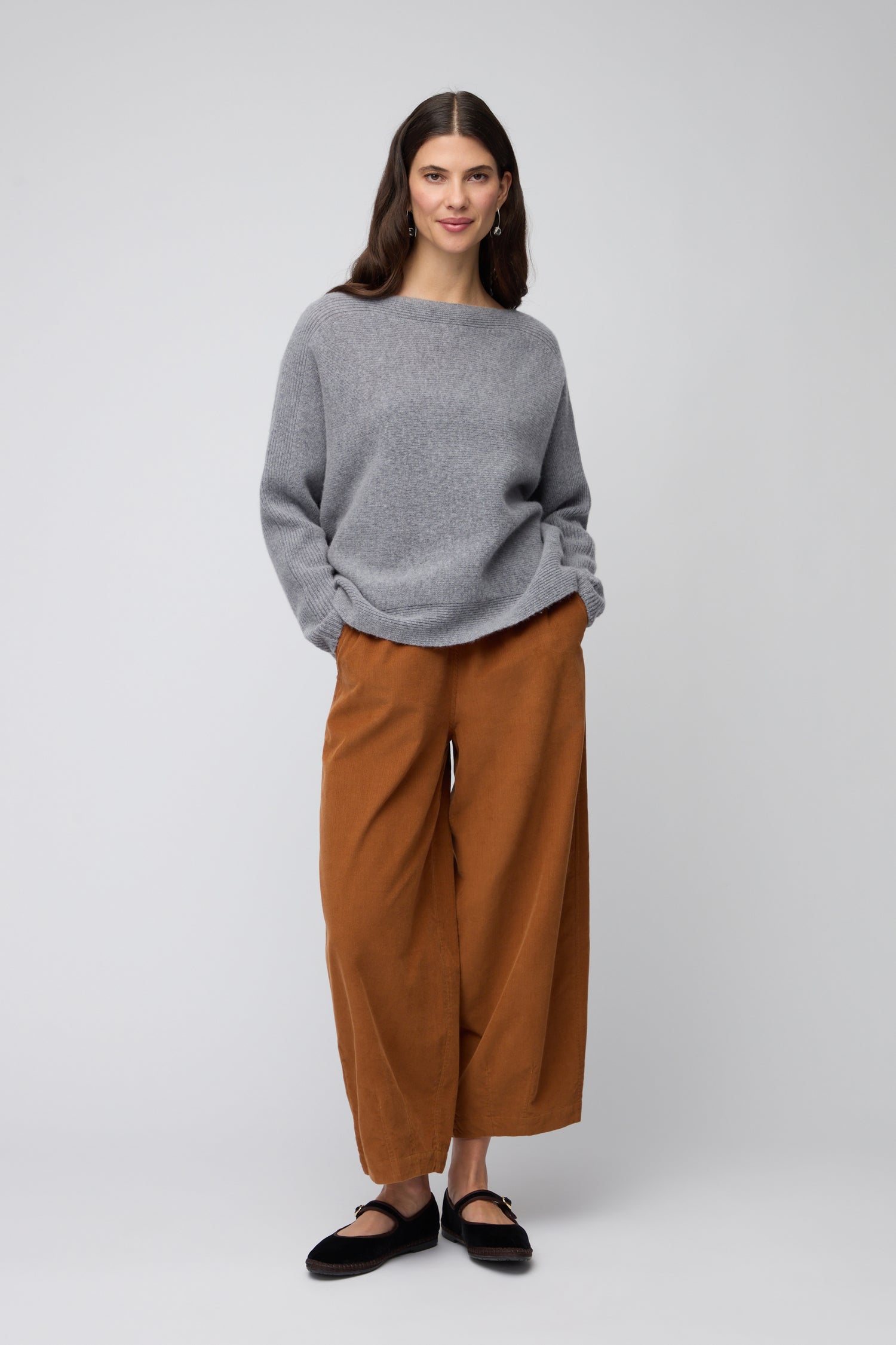 A woman faces forward against a plain light background, wearing the Merino Rib Detail Jumper—a lightweight knit grey sweater—paired with wide-legged brown pants and black shoes.