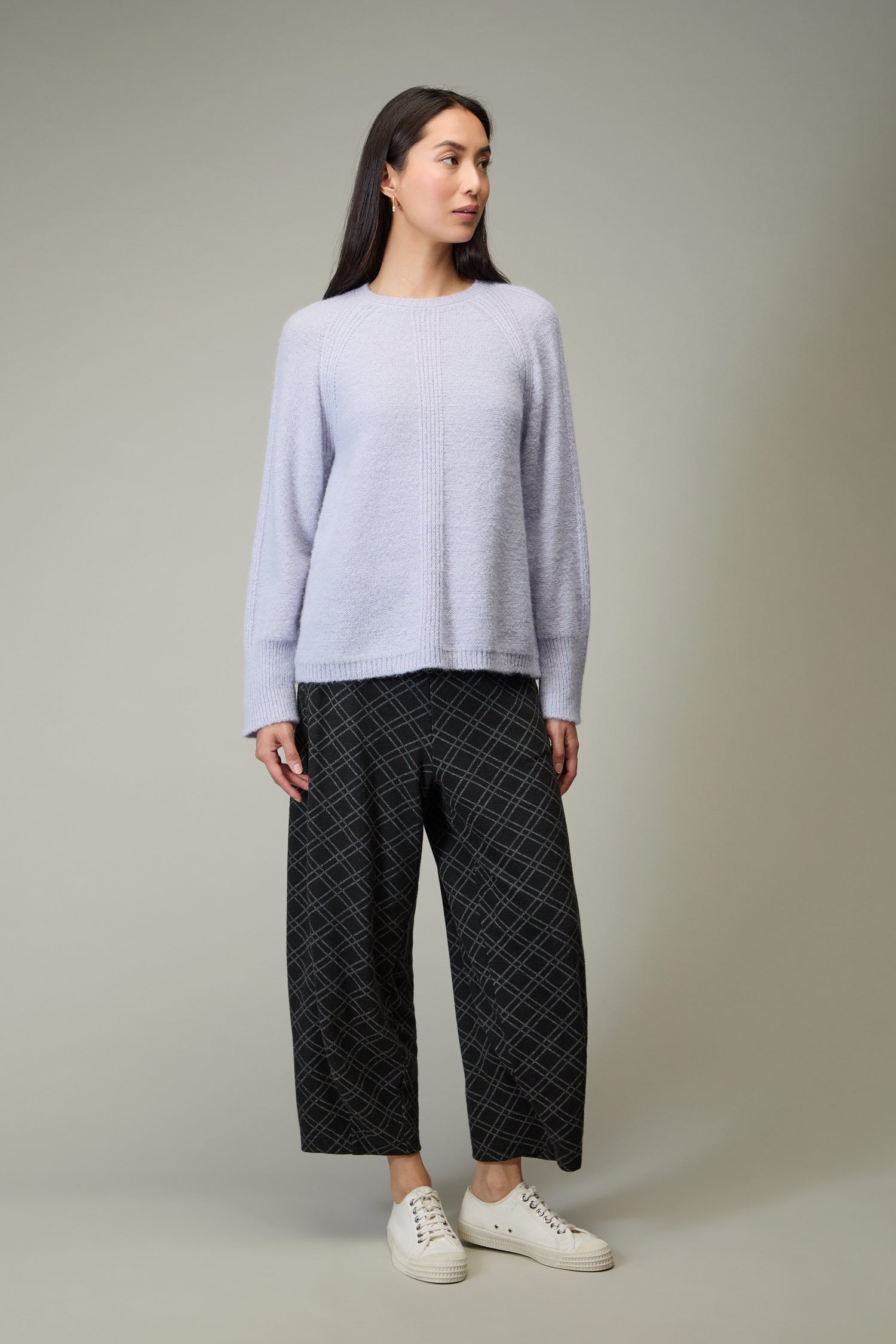 A woman stands against a plain background wearing the Alpaca Blend Seam Detail Jumper in light purple, black wide-leg grid patterned pants, and white sneakers.