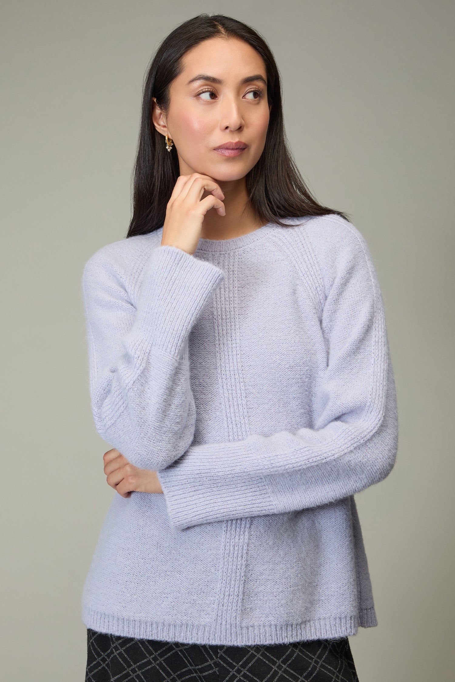 Woman with long dark hair wears the Alpaca Blend Seam Detail Jumper in light purple and a patterned dark skirt, standing against a plain gray background, looking left with her hand near her chin.