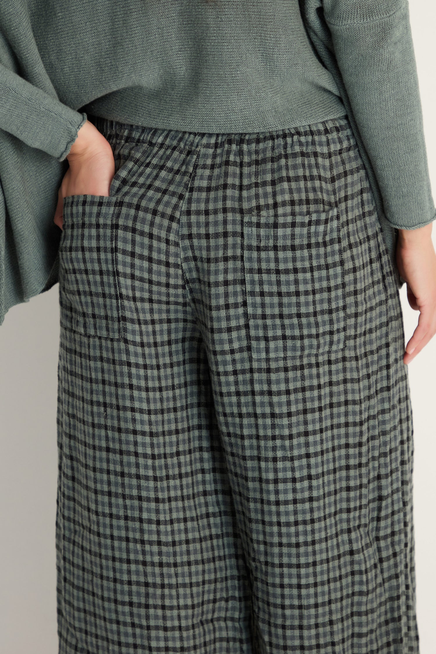 Model shown from the back waist down wearing Check Linen Bubble Trousers in a relaxed silhouette, paired with a textured green top and one hand in their pocket.