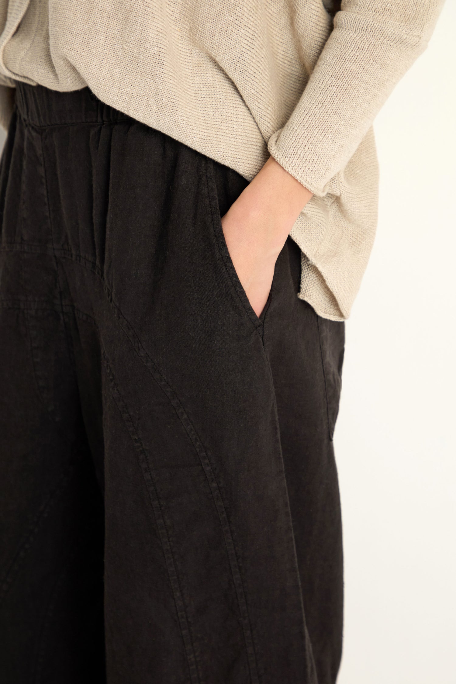 Model shown from mid-torso to mid-thigh against a light background, wearing Curved Seam Linen Trousers and a beige sweater, with one hand in the pants pocket.