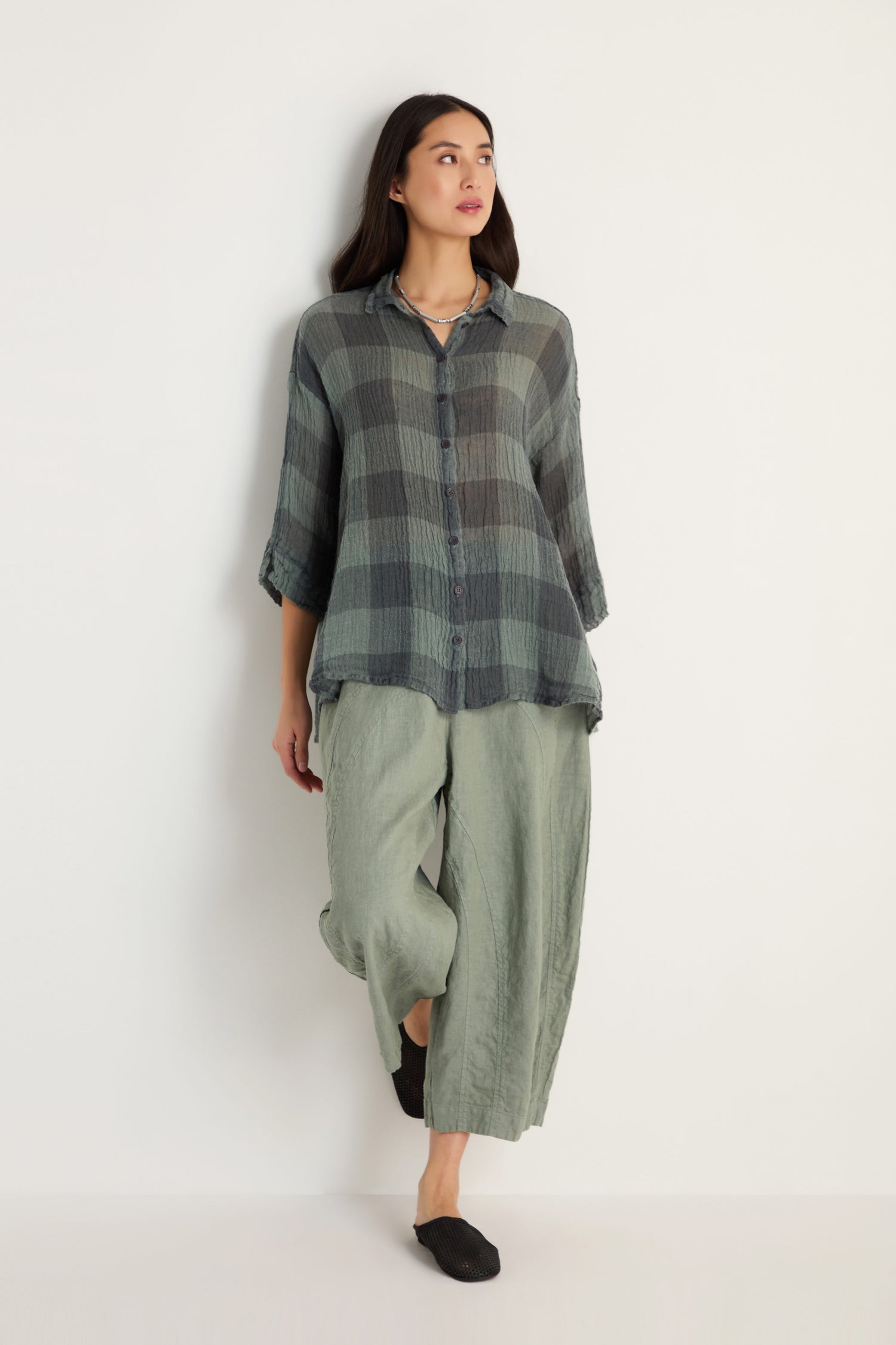 Against a white background, a woman wears the Large Check Sage Linen Swing Shirt in a relaxed silhouette, paired with wide-leg light green pants and black slip-on shoes.