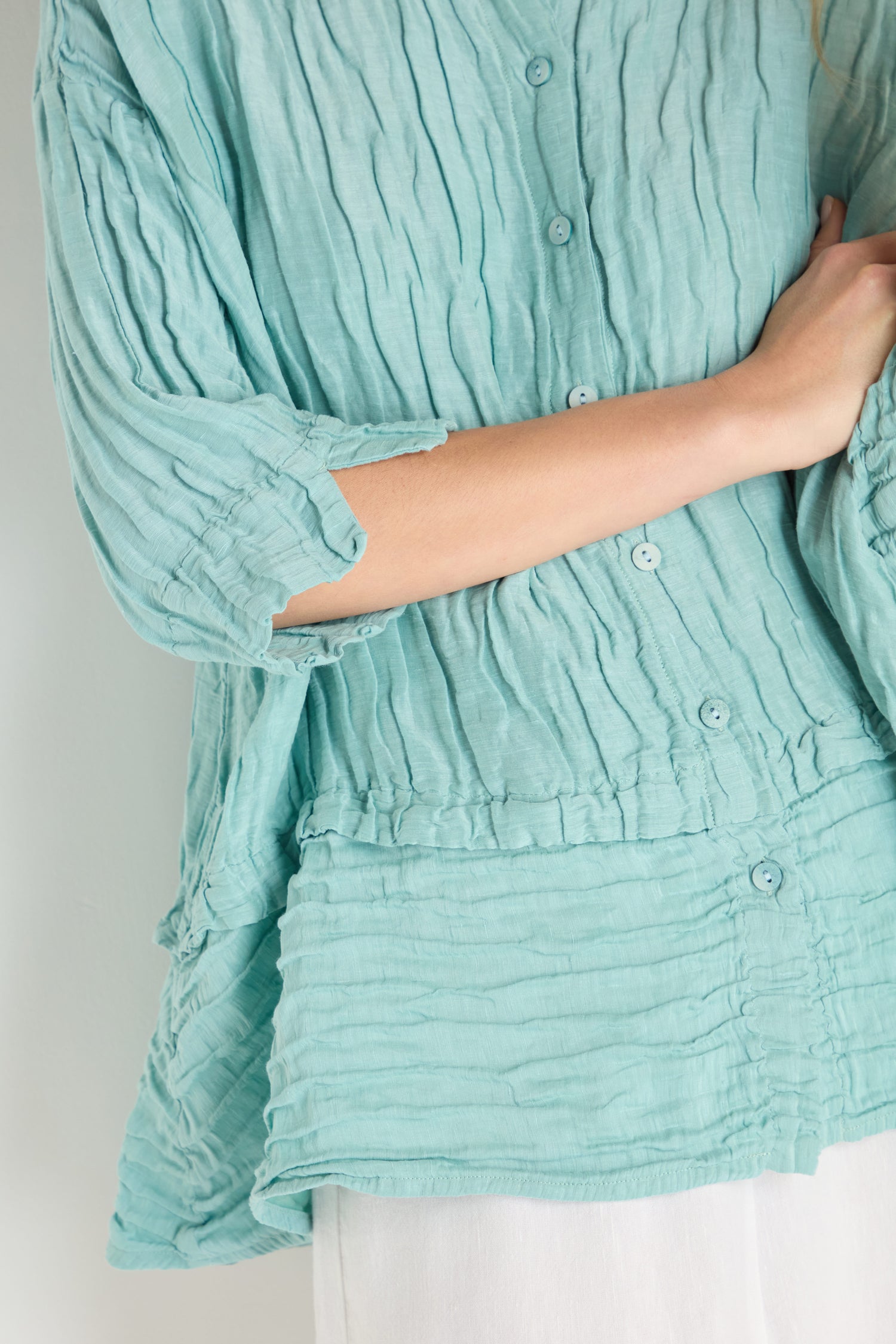 A person wears a light blue Panel Hem Silk Linen Shirt with three-quarter sleeves and white pants, standing with one arm crossed over their body.
