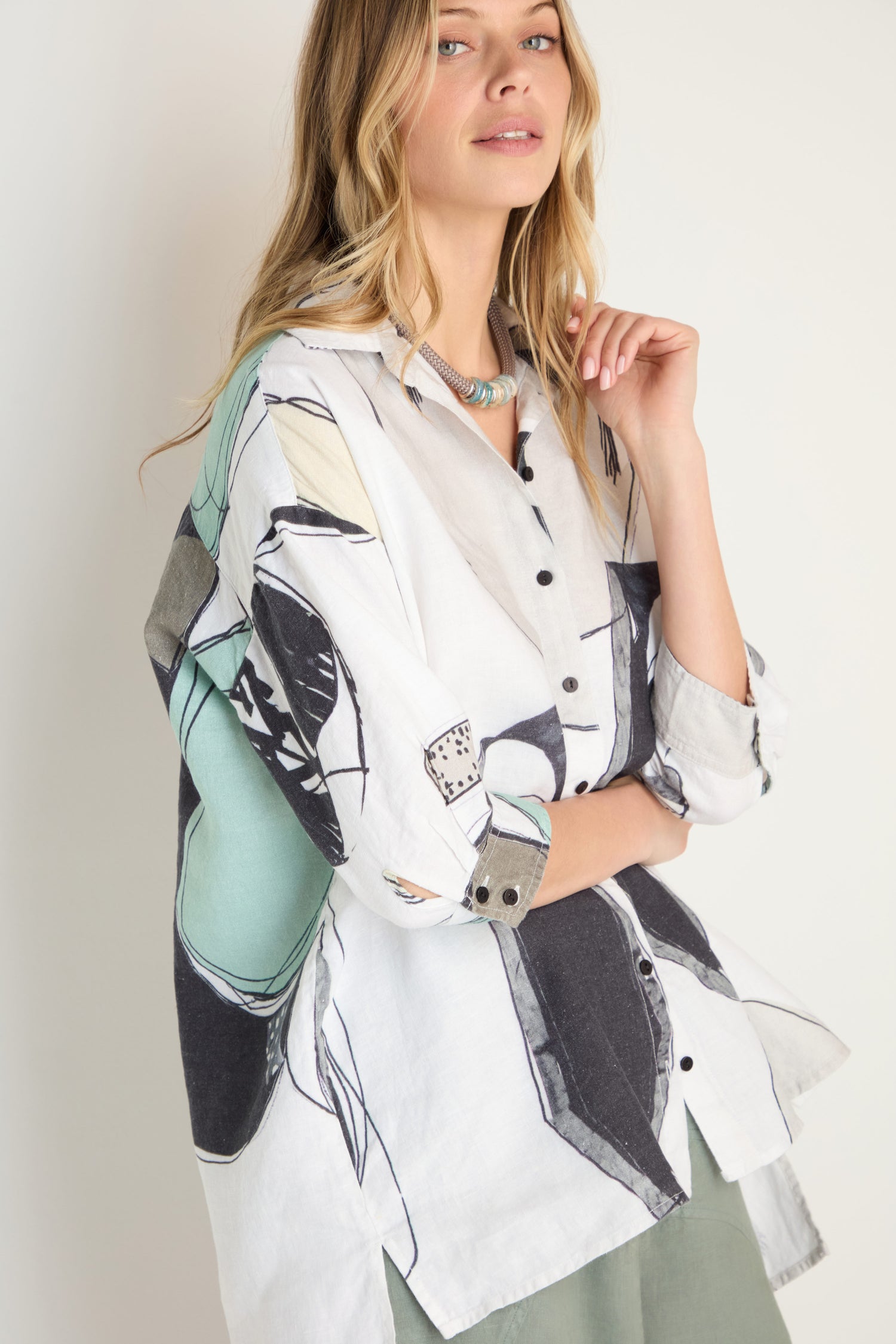 Woman wearing a long-sleeved, oversized Surreal Collage Print Linen Shirt by Grizas featuring abstract black, green, and beige patterns, standing against a plain background.