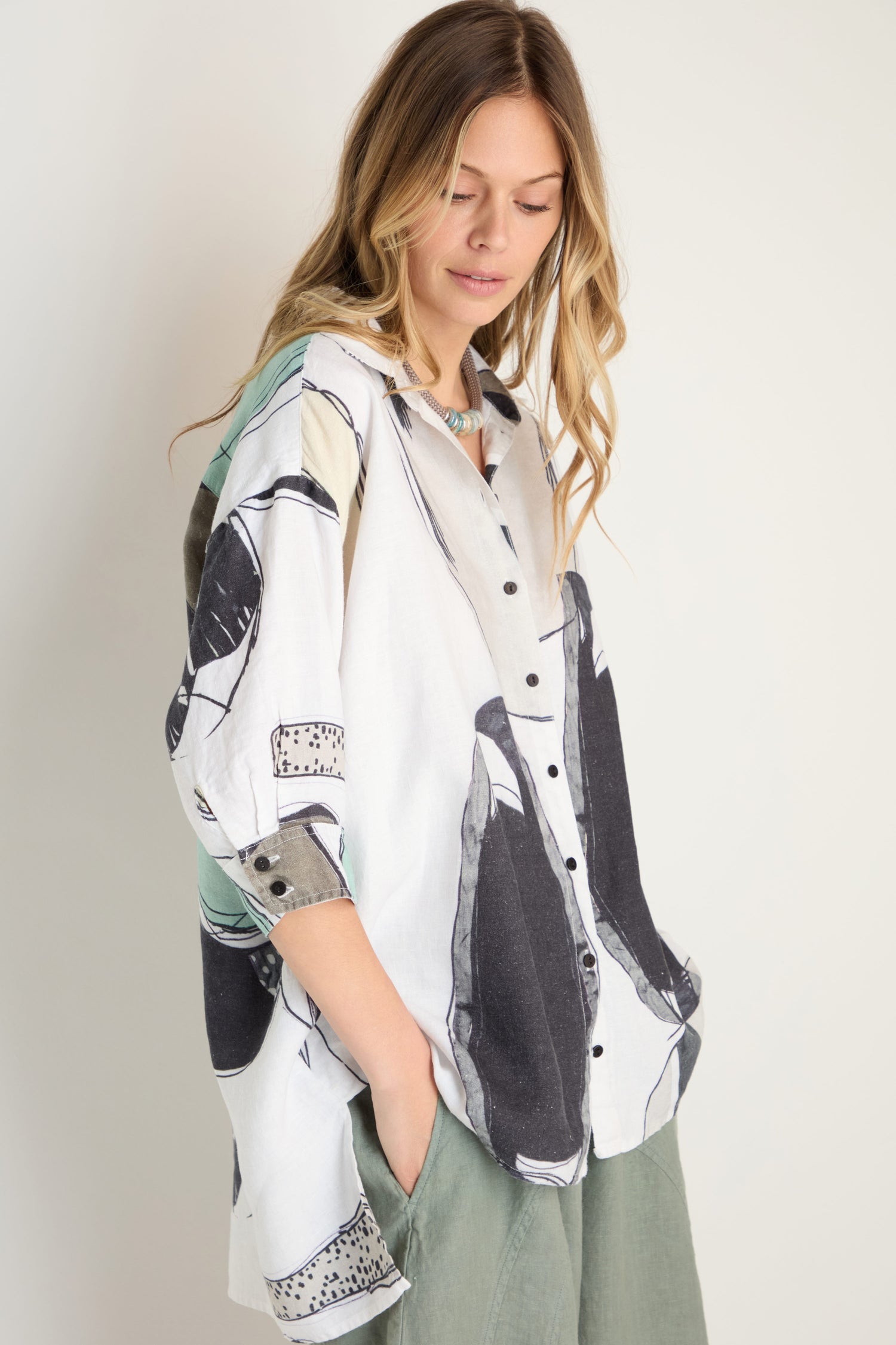 A woman with long blonde hair stands against a plain background, hands in her pockets, wearing the Surreal Collage Print Linen Shirt and green pants.