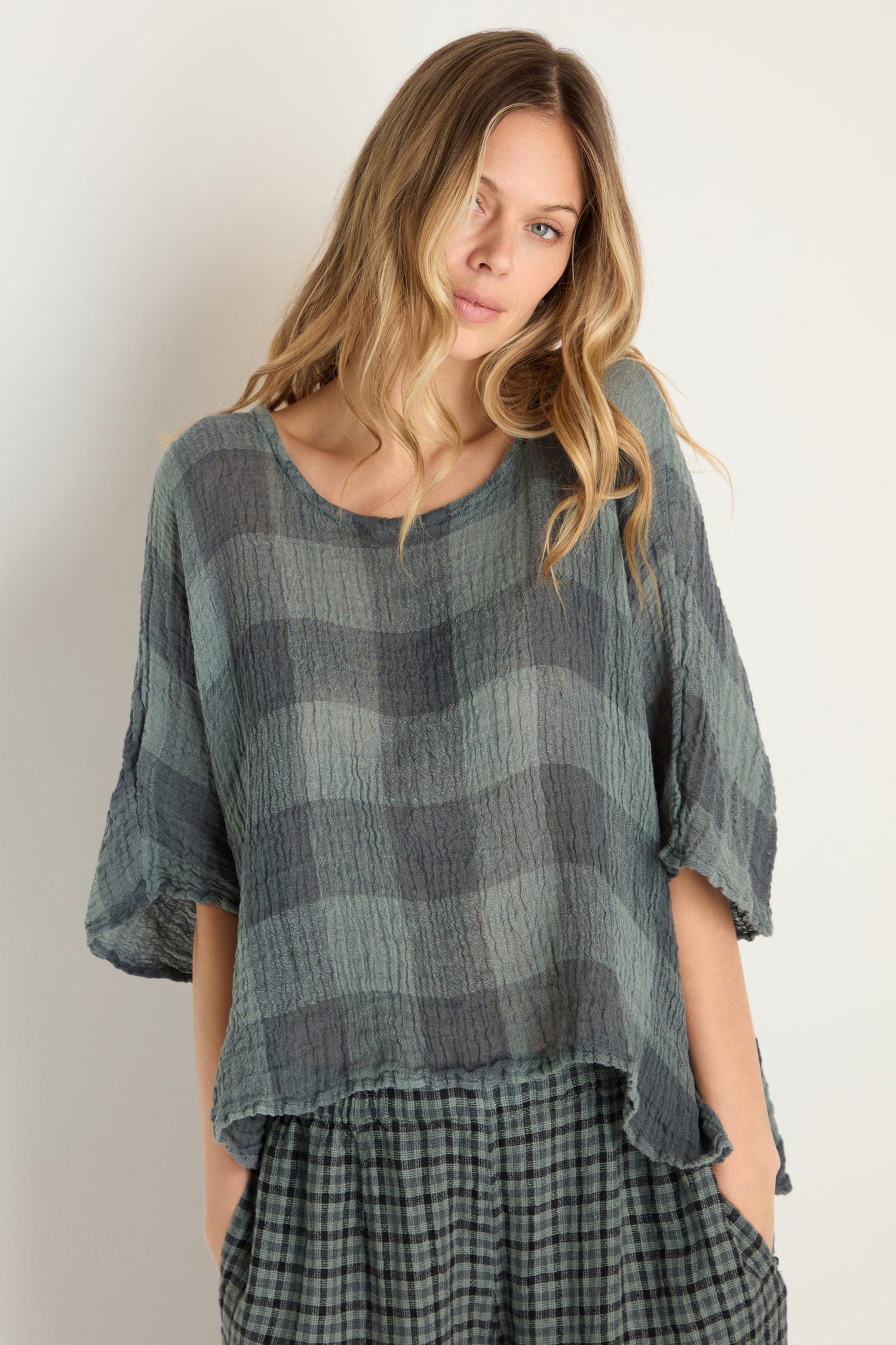 A woman stands against a light background wearing the Large Check Boxy Top— a loose, woven linen top with a blue-green large check pattern—paired with matching pants, her hands in her pockets.