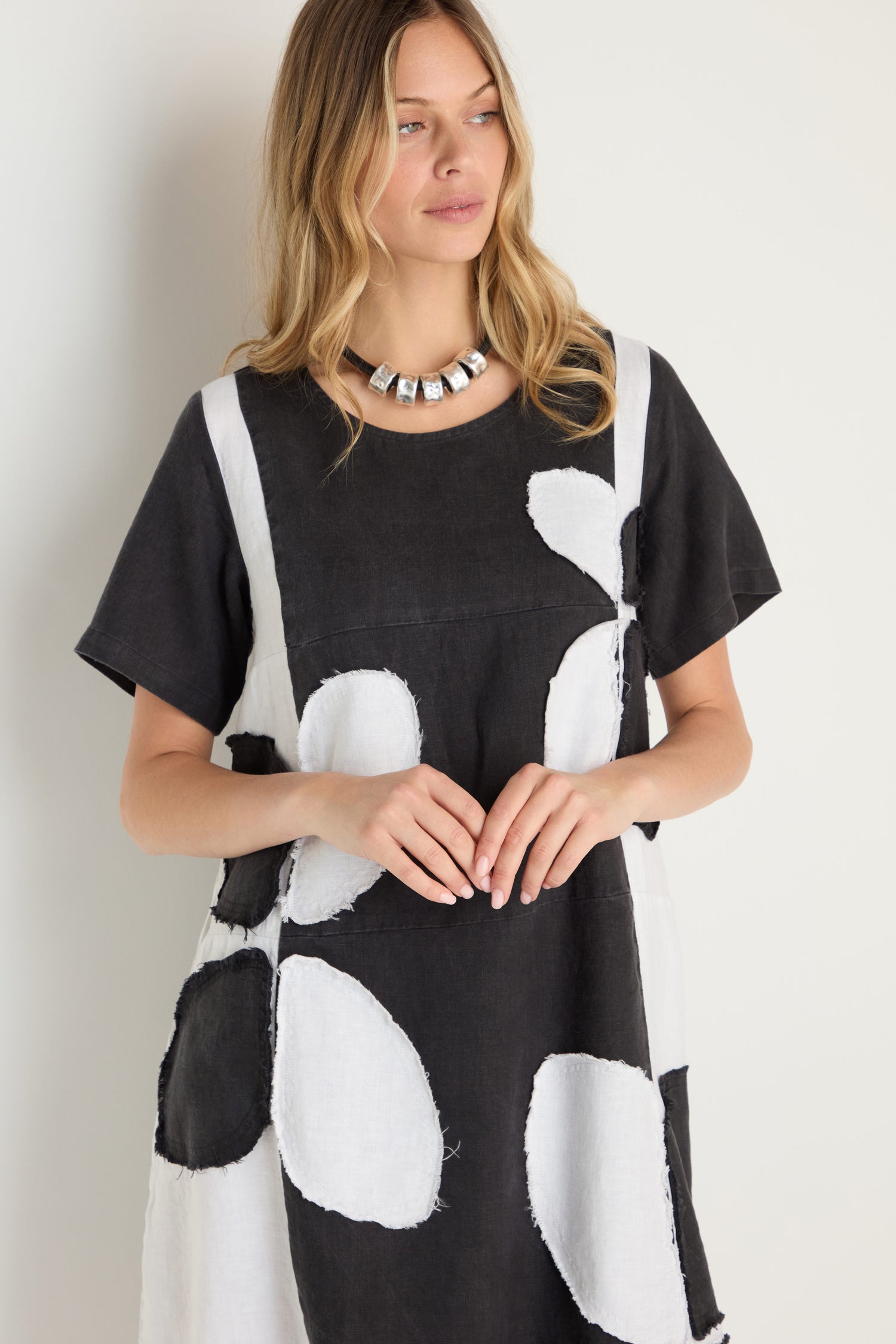 A woman with long blonde hair models the Quant Monochrome Linen Dress, featuring bold black and white abstract shapes, paired with a chunky silver necklace against a simple light backdrop.