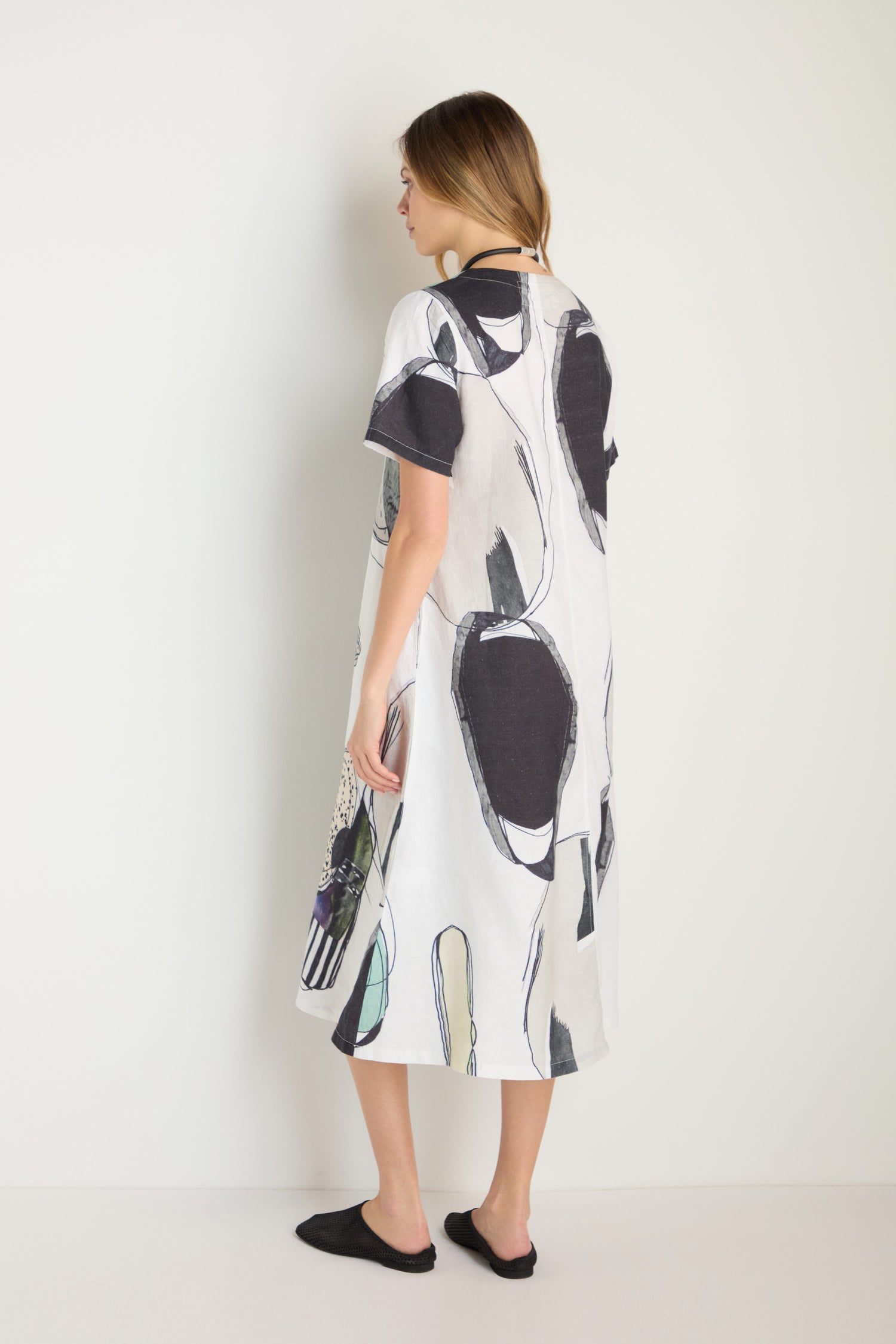 A woman stands against a white wall, wearing the Surreal Collage Print Linen Dress—a loose-fitting, short-sleeve linen piece with an abstract black and white print—paired with black slip-on shoes.