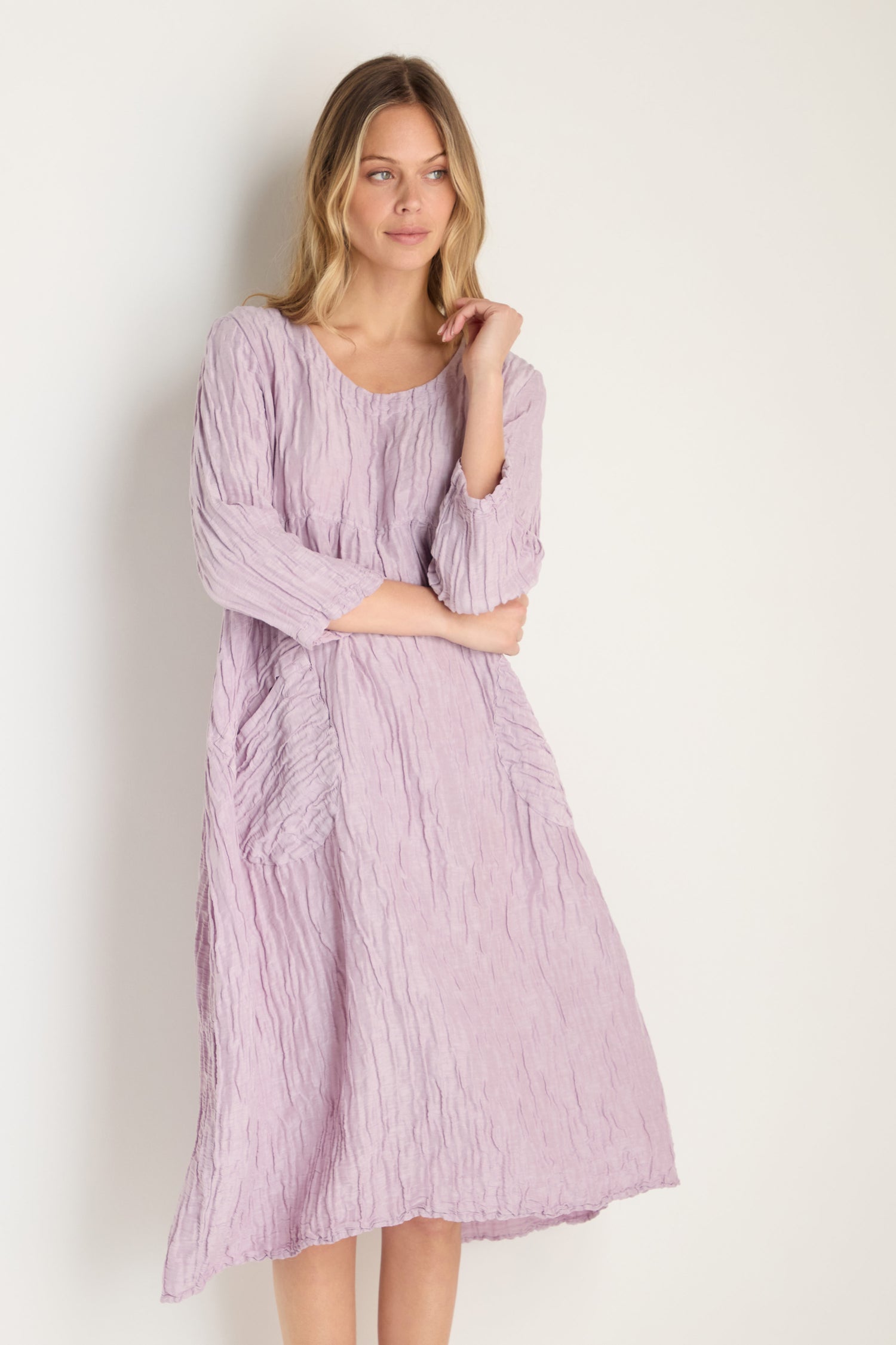 A woman stands against a white background, wearing the Silk Linen Lilac Patch Pocket Dress—loose-fitting with long sleeves and patch pockets—gazing slightly to the side.
