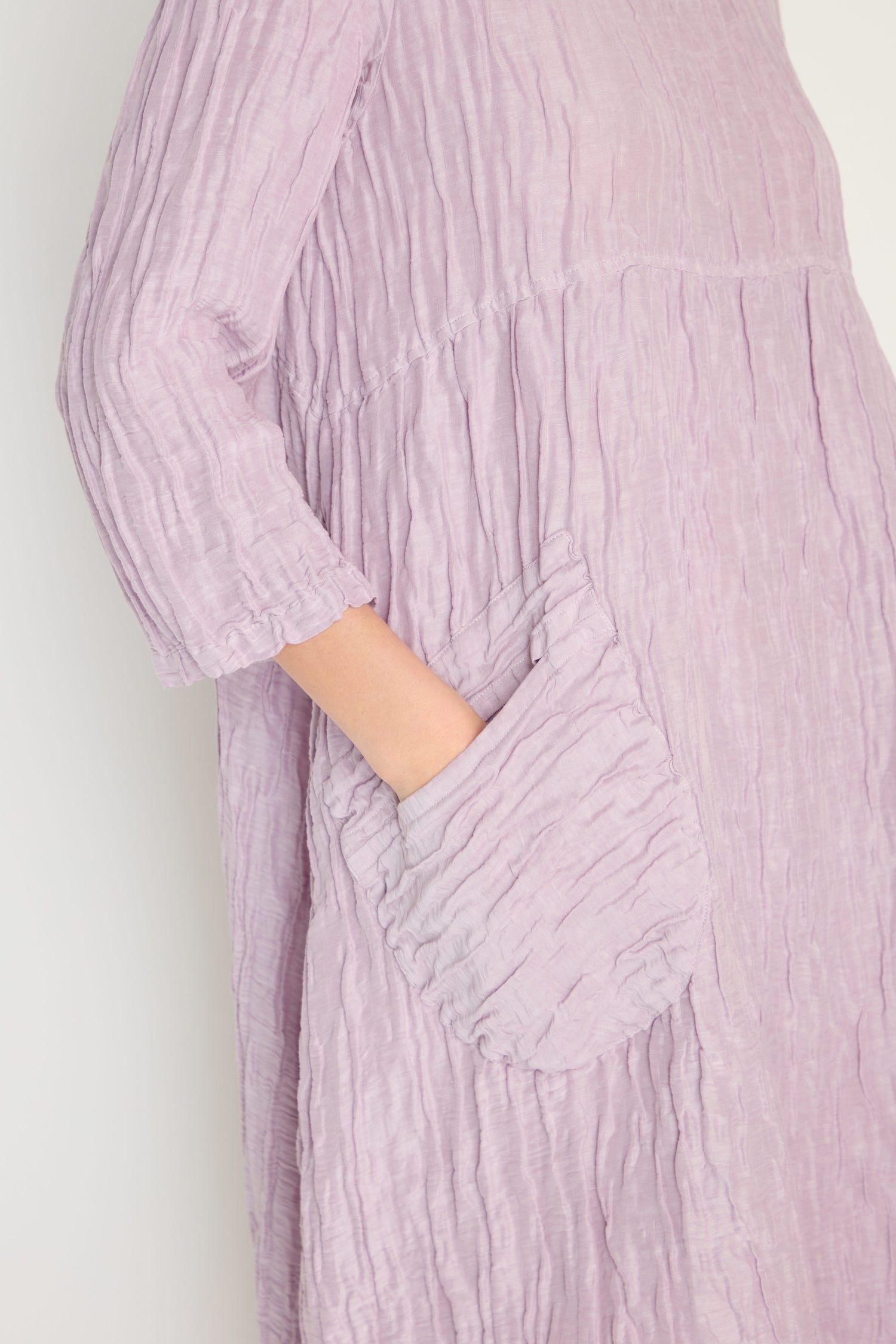 A person wears the Silk Linen Lilac Patch Pocket Dress, a light purple textured dress with three-quarter sleeves, and puts their hand in a side pocket.