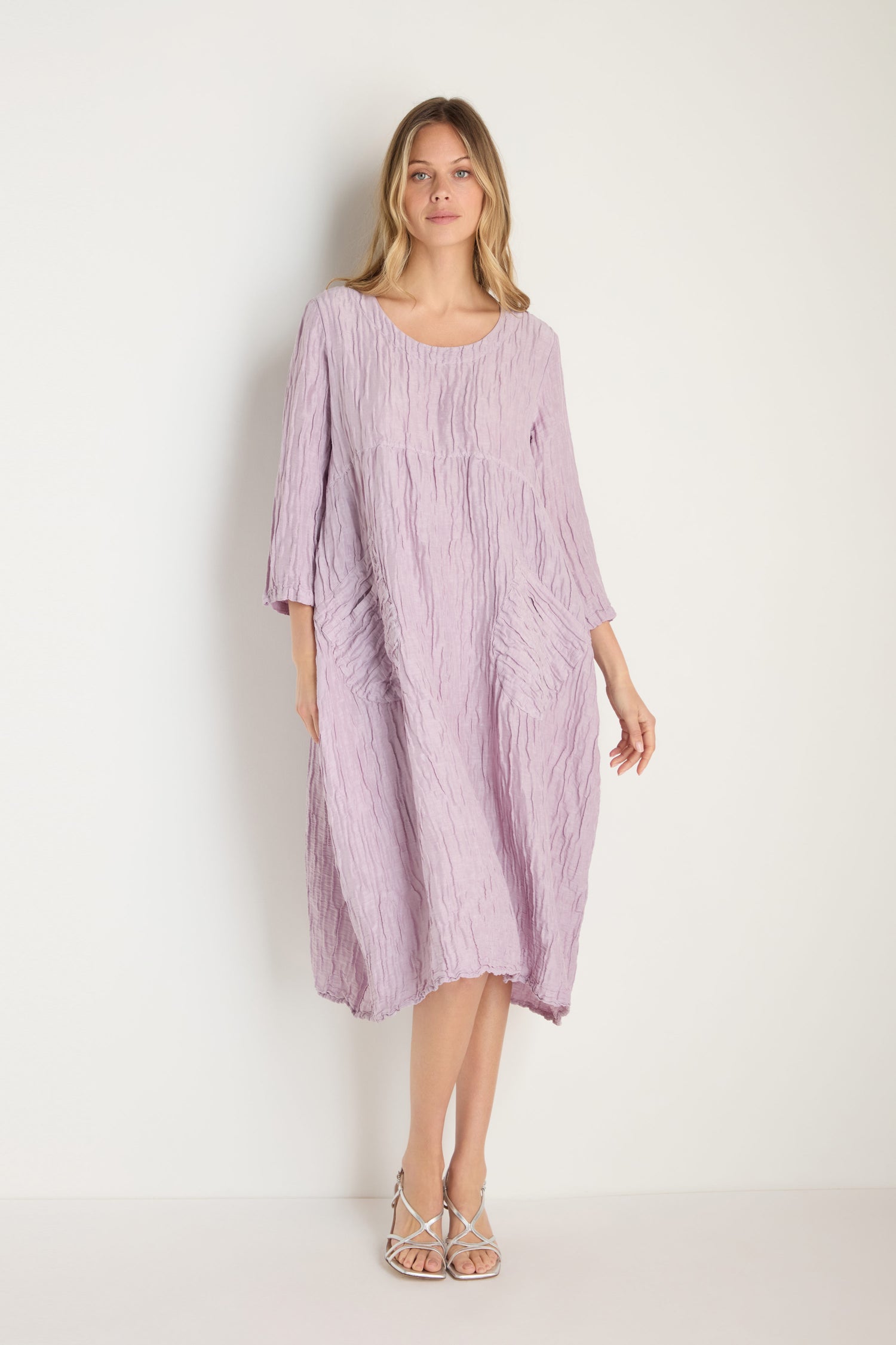 A woman stands against a plain background wearing the Silk Linen Lilac Patch Pocket Dress—a loose, textured dress with three-quarter sleeves and side pockets—paired with strappy sandals.