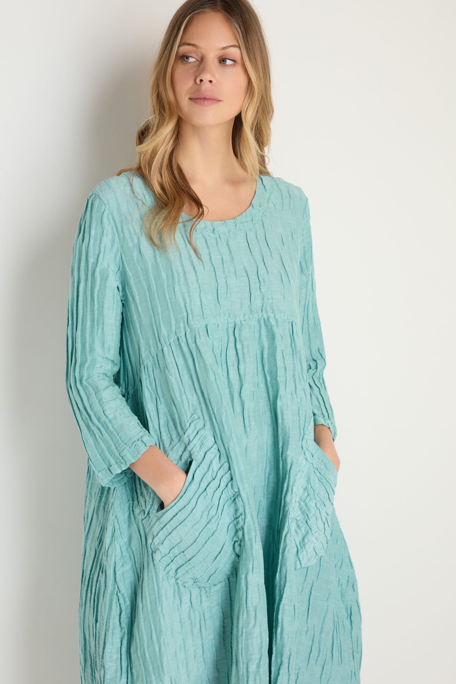 A woman stands against a plain light background wearing the Silk Linen Mint Patch Pocket Dress, a textured, light teal long-sleeve dress featuring large front pockets.