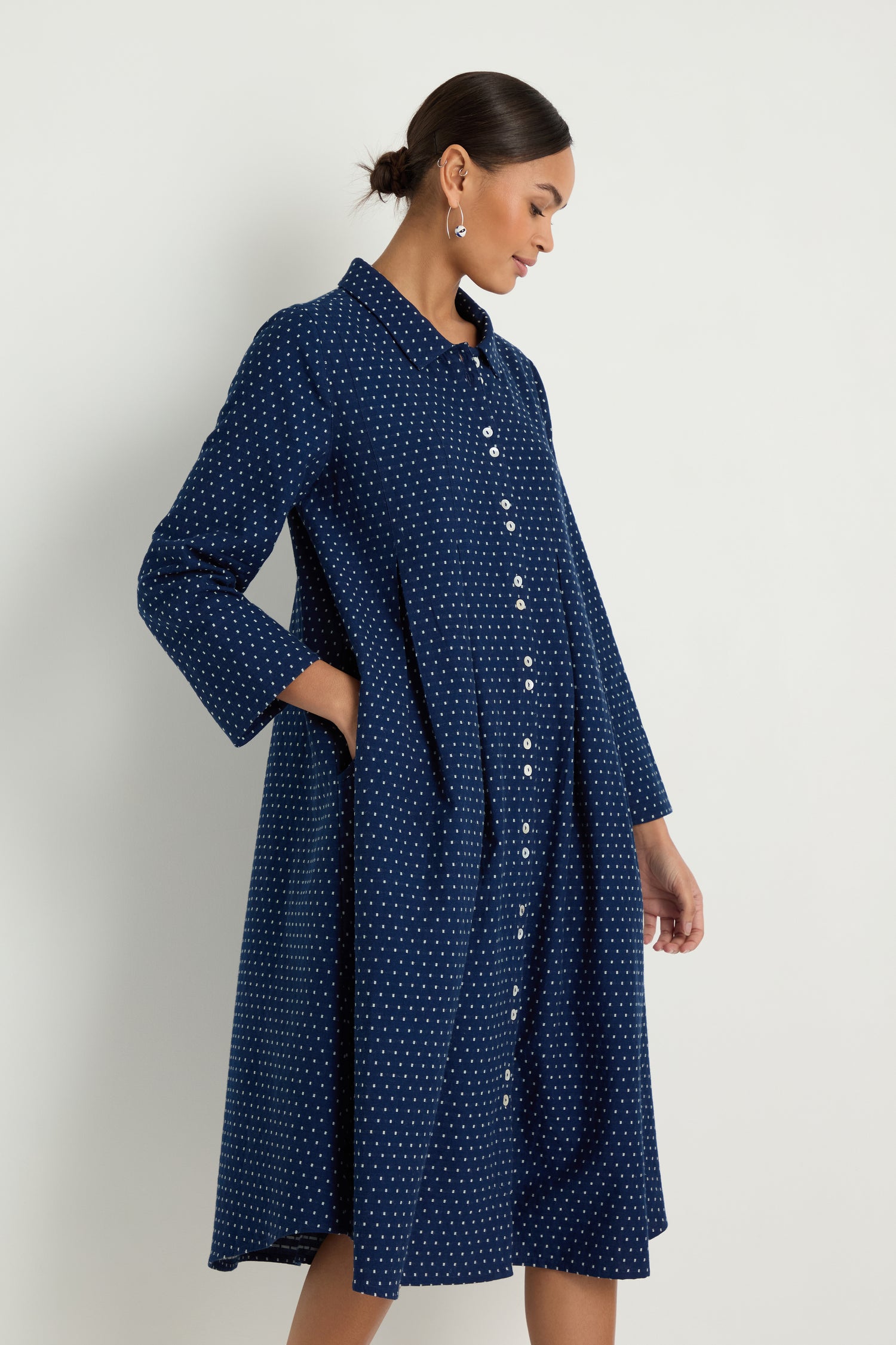 A woman models the Indigo Spot Cotton Shirt Dress, a long-sleeved, knee-length navy cotton dress with white polka dots and front buttons, highlighting its relaxed fit and subtle A-line silhouette against a white background.