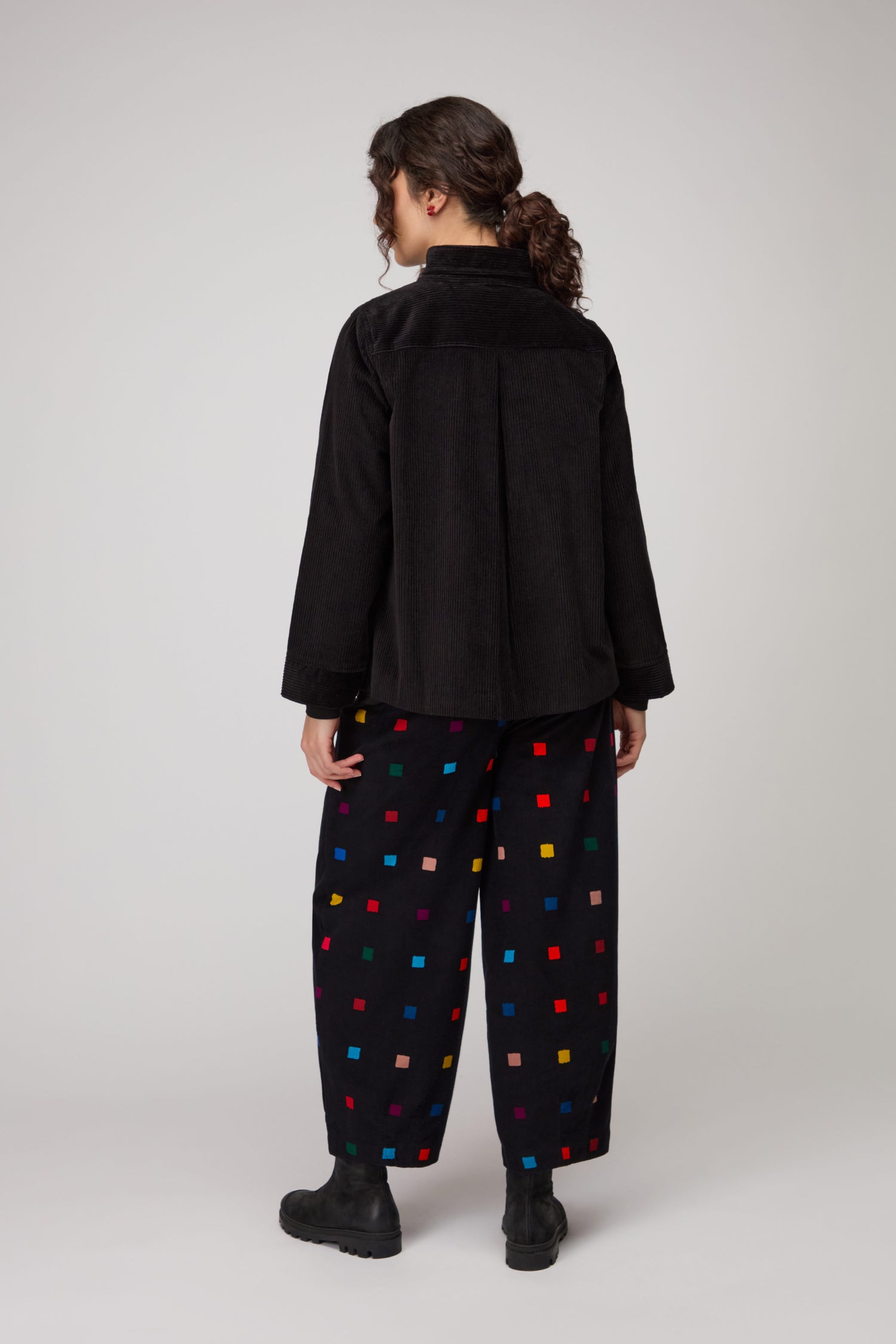 Person standing facing away, wearing a Jumbo Cord Jacket and loose black pants with colorful square patterns, against a plain light background.