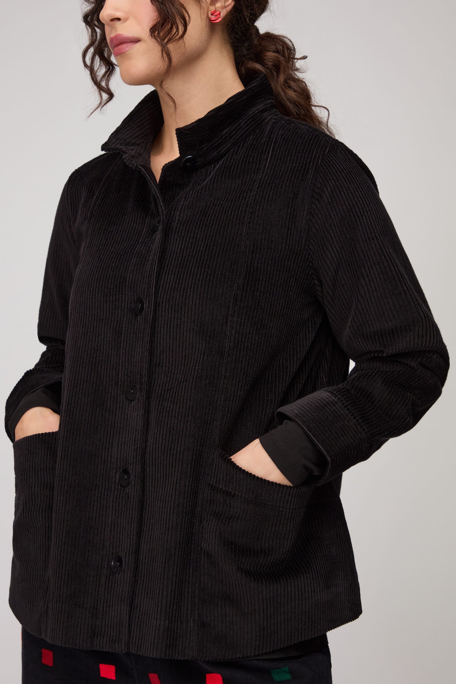 A woman stands against a plain background with her hands in the front pockets of a black Jumbo Cord Jacket.