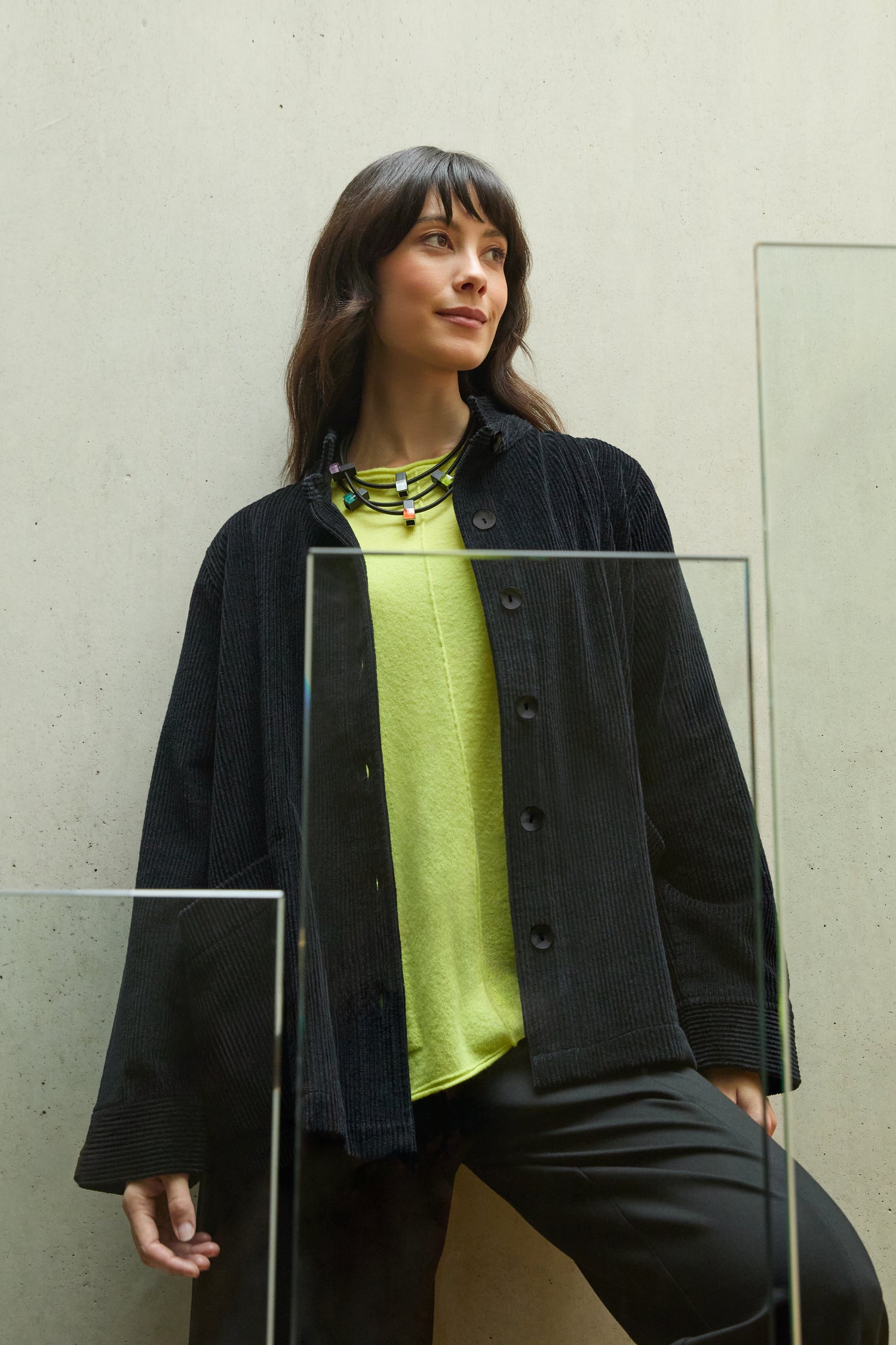 Woman standing indoors against a light-colored wall, wearing the Jumbo Cord Jacket over a lime green top and black pants, paired with a multicolored necklace.