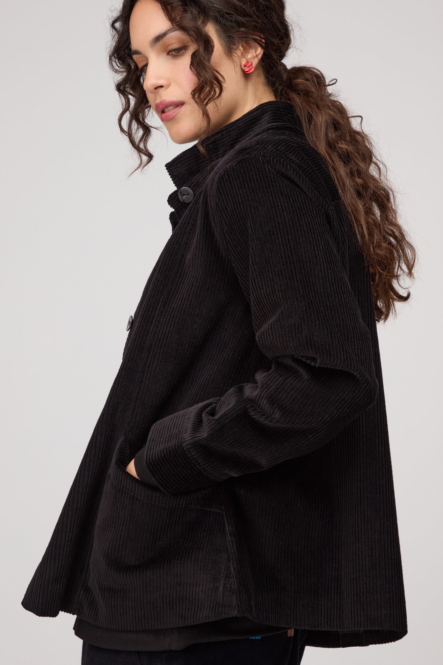 A woman with curly brown hair poses in profile against a plain background, wearing the black Jumbo Cord Jacket with one hand in her pocket.