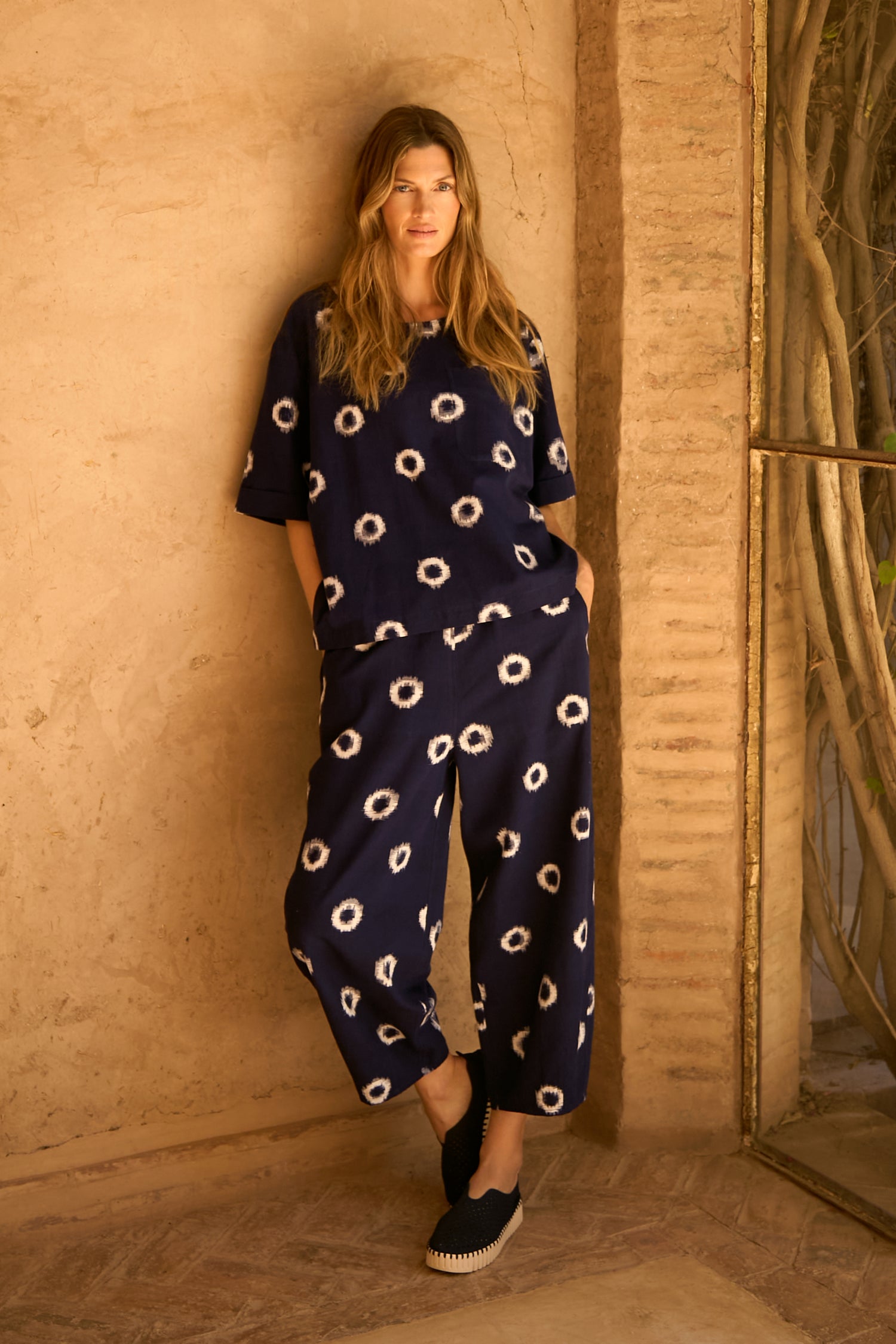 A woman stands against a beige textured wall, wearing Ikat Circles Cotton Bubble Trousers—loose-fitting navy pants with a white circular pattern—paired with a matching top and black slip-on shoes.