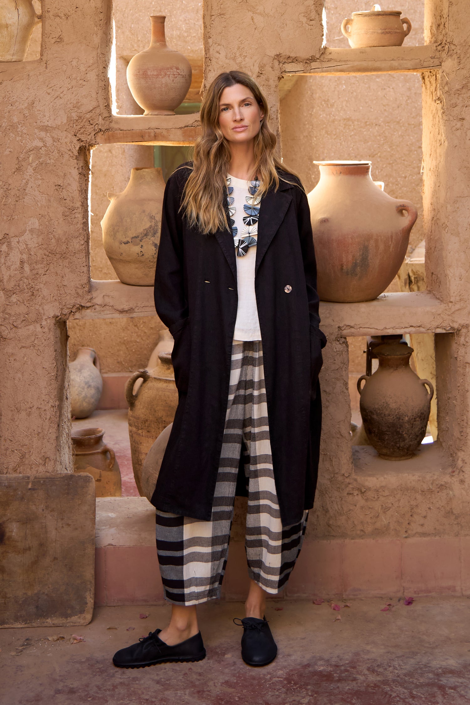 A woman stands by a rustic wall with pottery, modeling the GRP5167 look: a black coat, white top, Monochrome Crinkle Check Bubble Trouser, and black shoes.