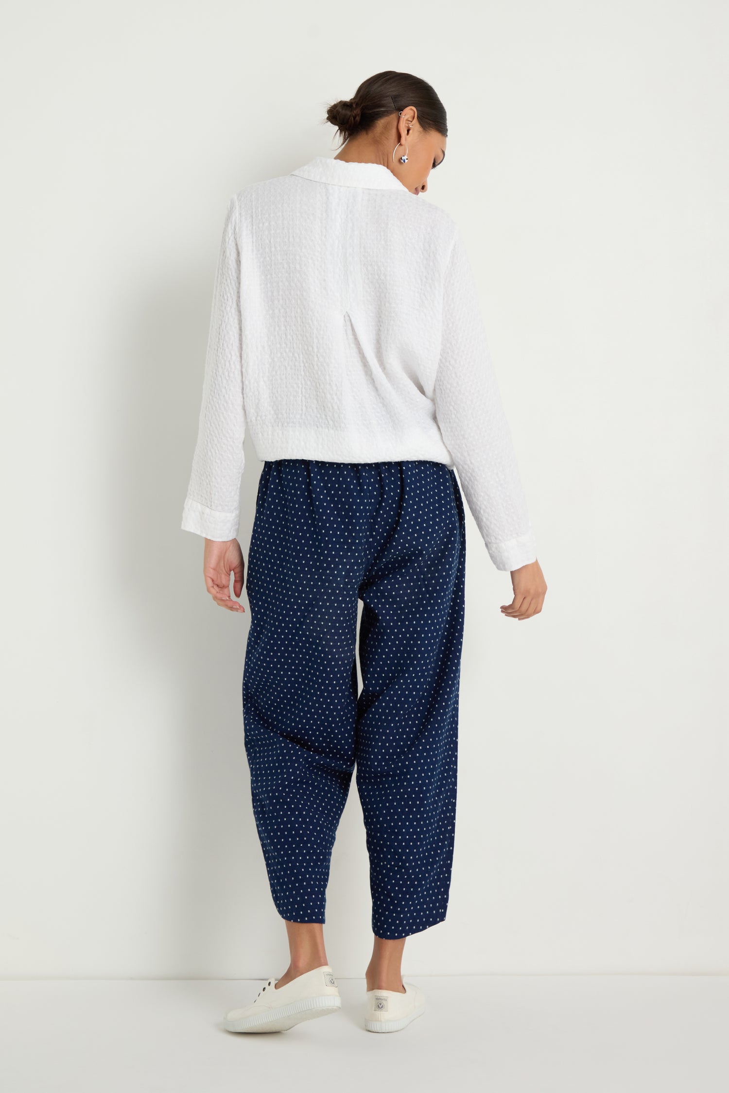 A woman stands against a white wall, facing away, wearing a white textured long-sleeve top and the Indigo Spot Cotton Bubble Trouser with an elasticated waist and cropped bubble silhouette, paired with white sneakers.
