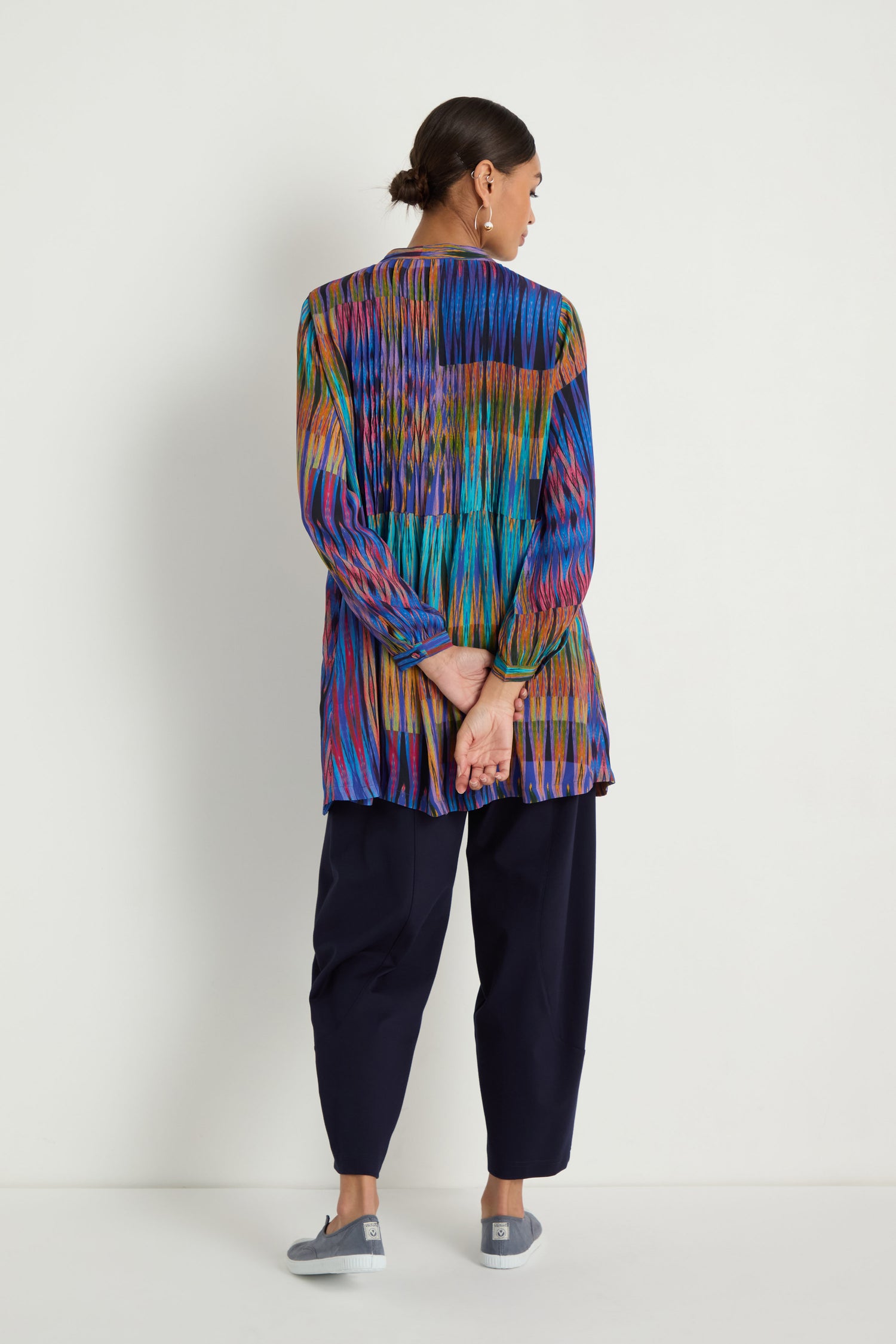 A person faces away from the camera, wearing the Vivid Ikat Patchwork Top with long sleeves, dark loose pants, gray slip-on shoes, and has their hands clasped behind their back.