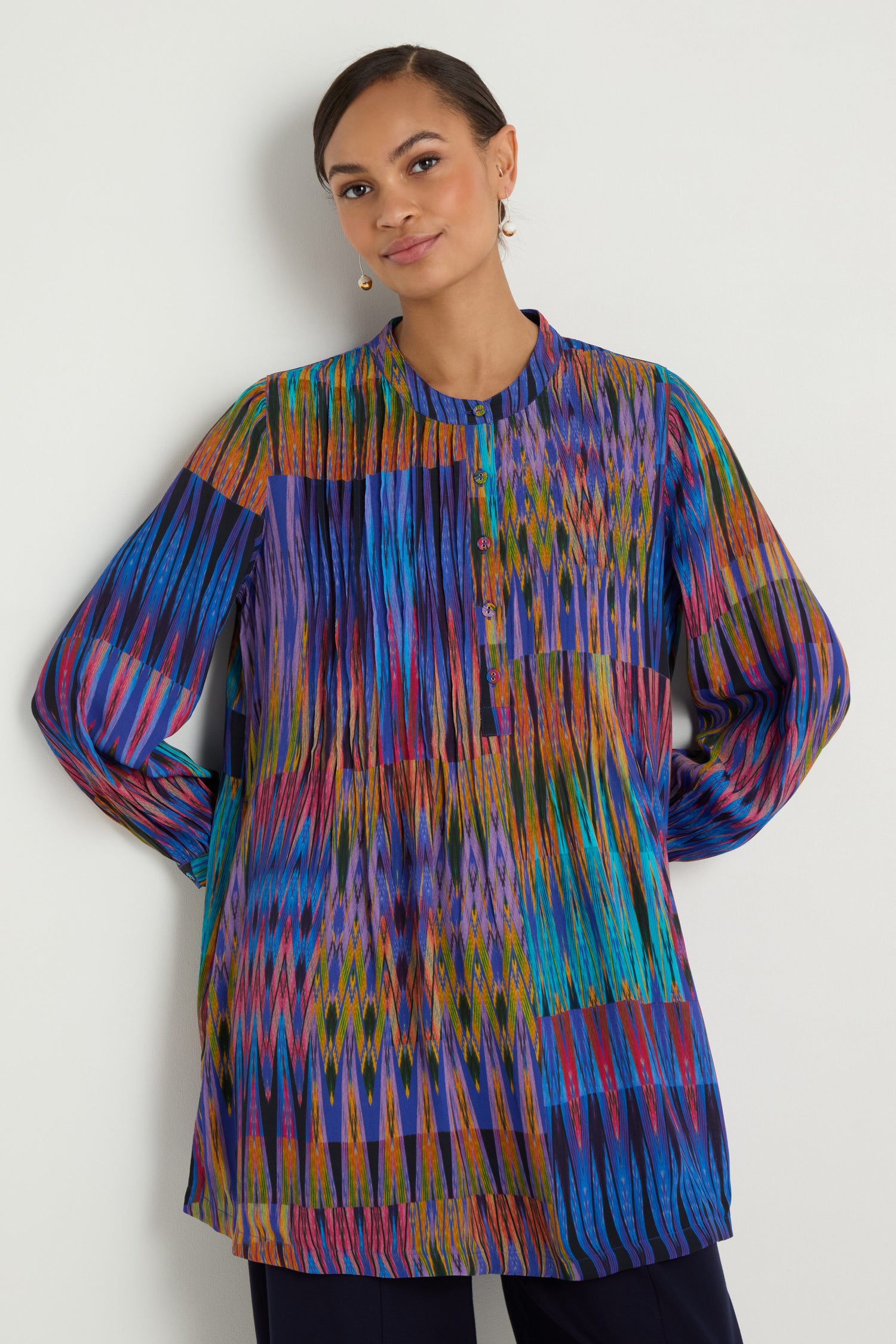 A woman stands against a plain wall wearing the Vivid Ikat Patchwork Top, a long-sleeved tunic featuring a colorful print with blue, purple, red, and yellow tones.